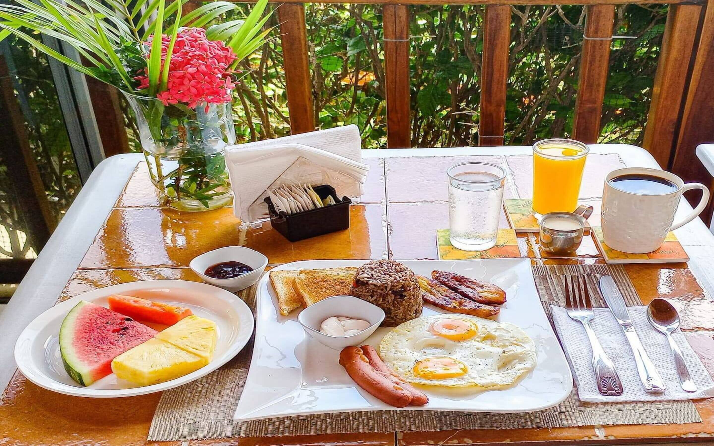 A breakfast table with plates of toast, eggs, sausages, and fruit. There is a coffee cup, a glass of orange juice, and a vase with flowers.