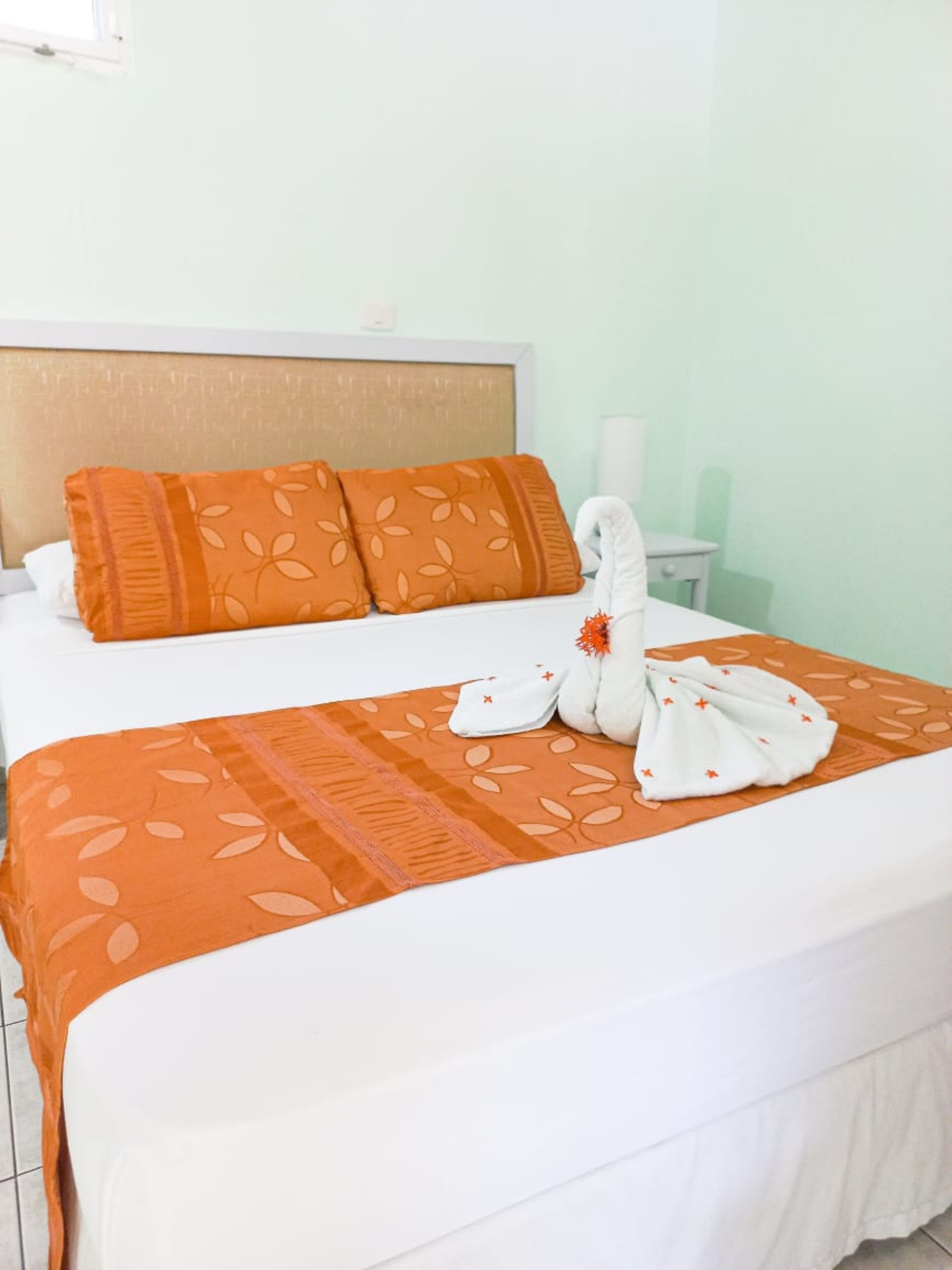 A neatly made bed with a white duvet, orange patterned pillows, and a towel arrangement on the bed.