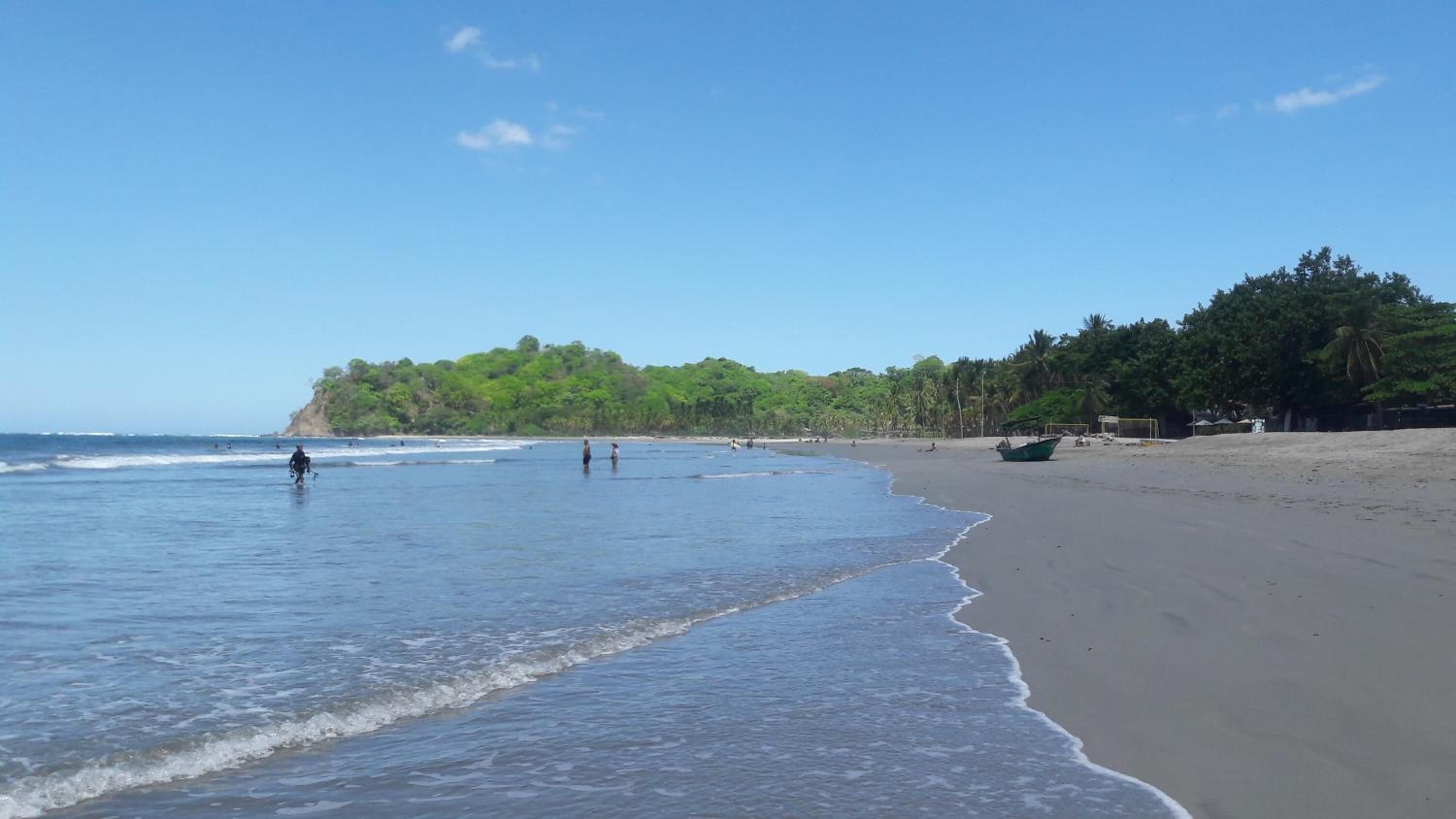 A beach with a boat on the sand, a person in the water, and a forested hill in the background.