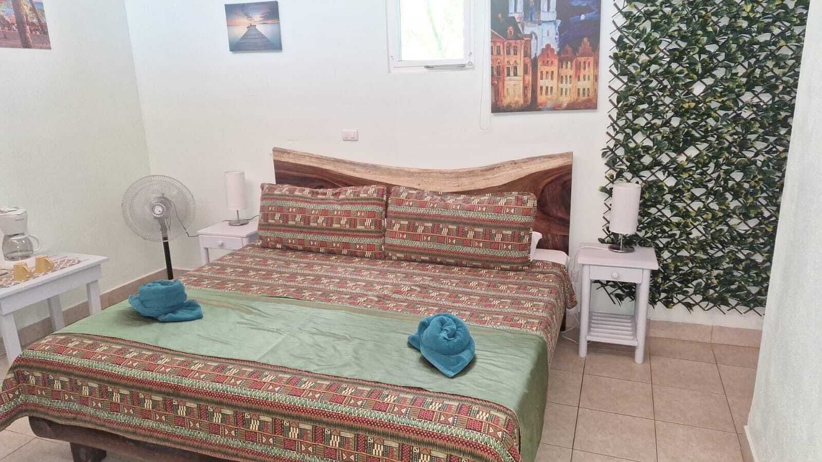A room with two beds, a fan, a small table with a lamp, and a wall with a leafy pattern.