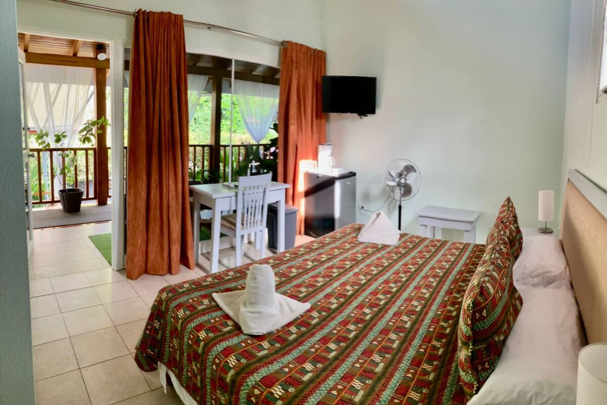 Habitación de hotel con cama, escritorio, silla, televisor y balcón con cortinas.