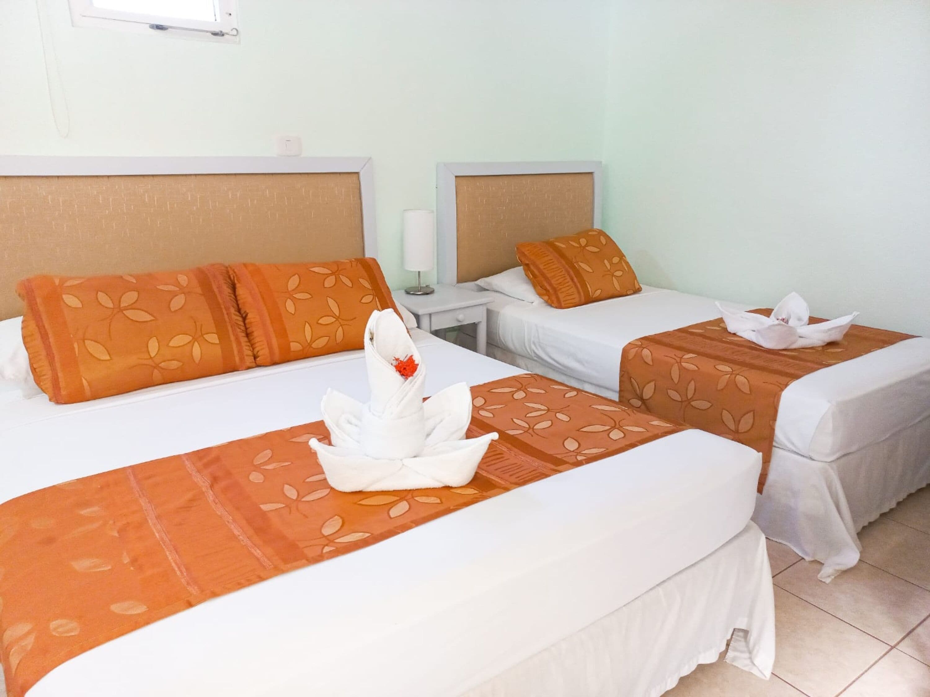 A hotel room with two single beds, each with an orange floral bedspread and matching pillows. A white towel is folded on the bed, and a white flower is placed on the pillow.