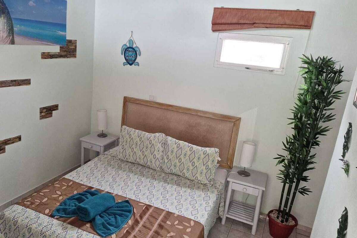 Un dormitorio con cama, mesitas de noche, ventana, un adorno en la pared y una planta en maceta.