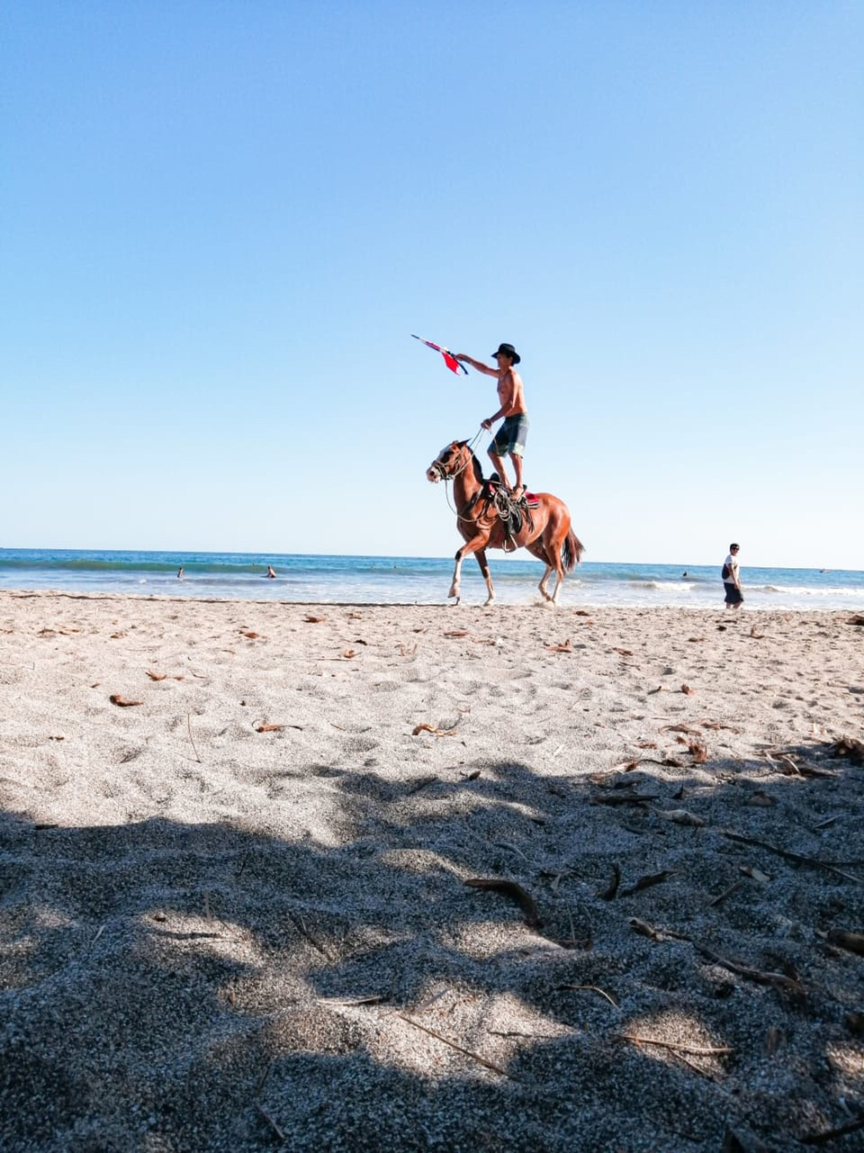 Une scène de plage où l’on voit une personne à cheval et une autre personne qui marche au loin.