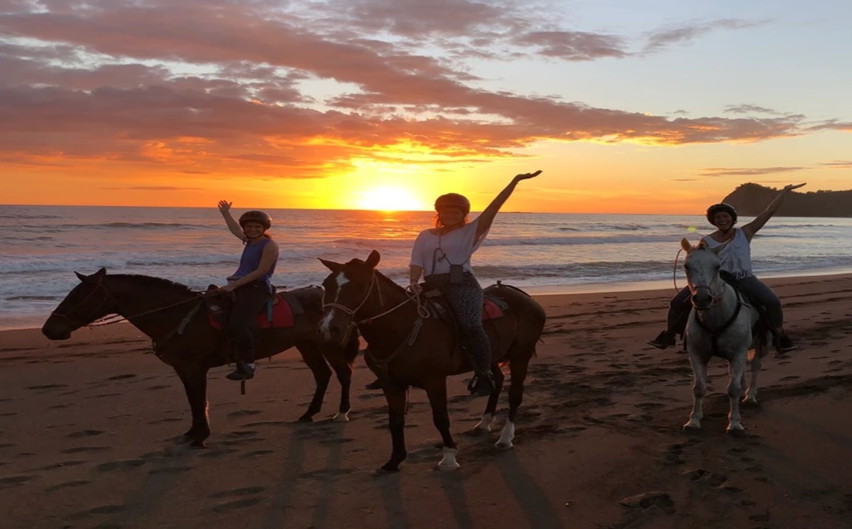 Three people riding horses on a beach at sunset.