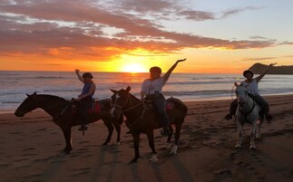 Three people riding horses on a beach at sunset.