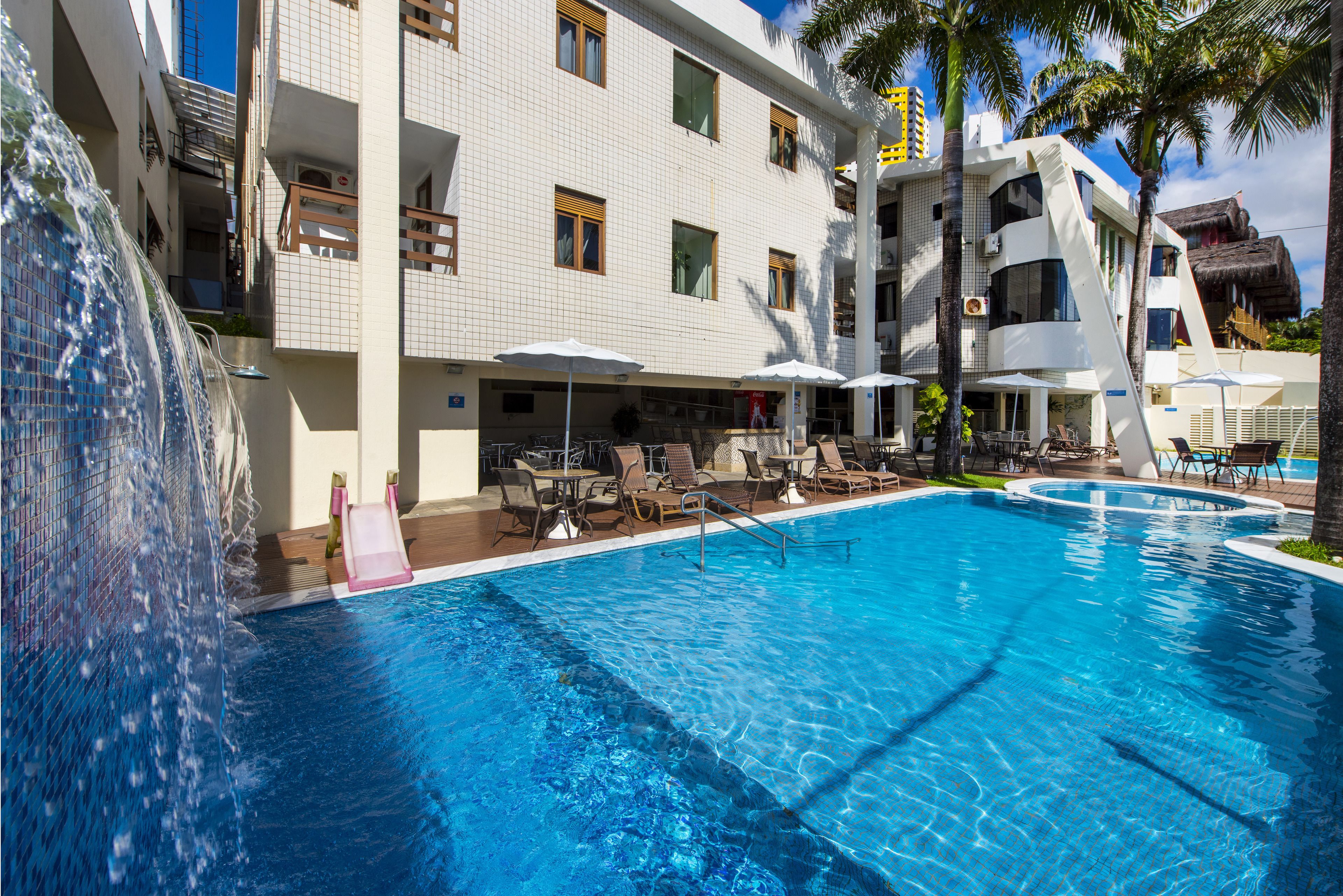 Un espace piscine d’hôtel avec une piscine à l’eau cristalline, des chaises longues, des parasols et un élément décoratif aquatique.