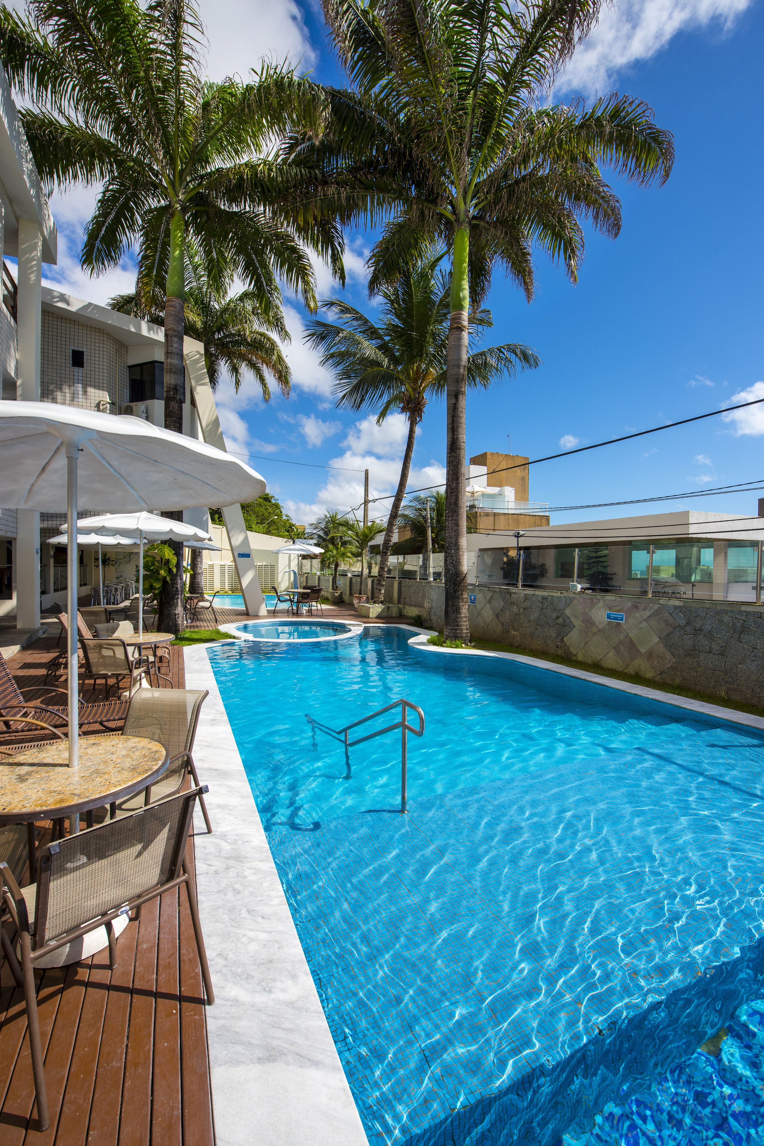 A swimming pool with lounge chairs and umbrellas, surrounded by palm trees and a clear blue sky.
