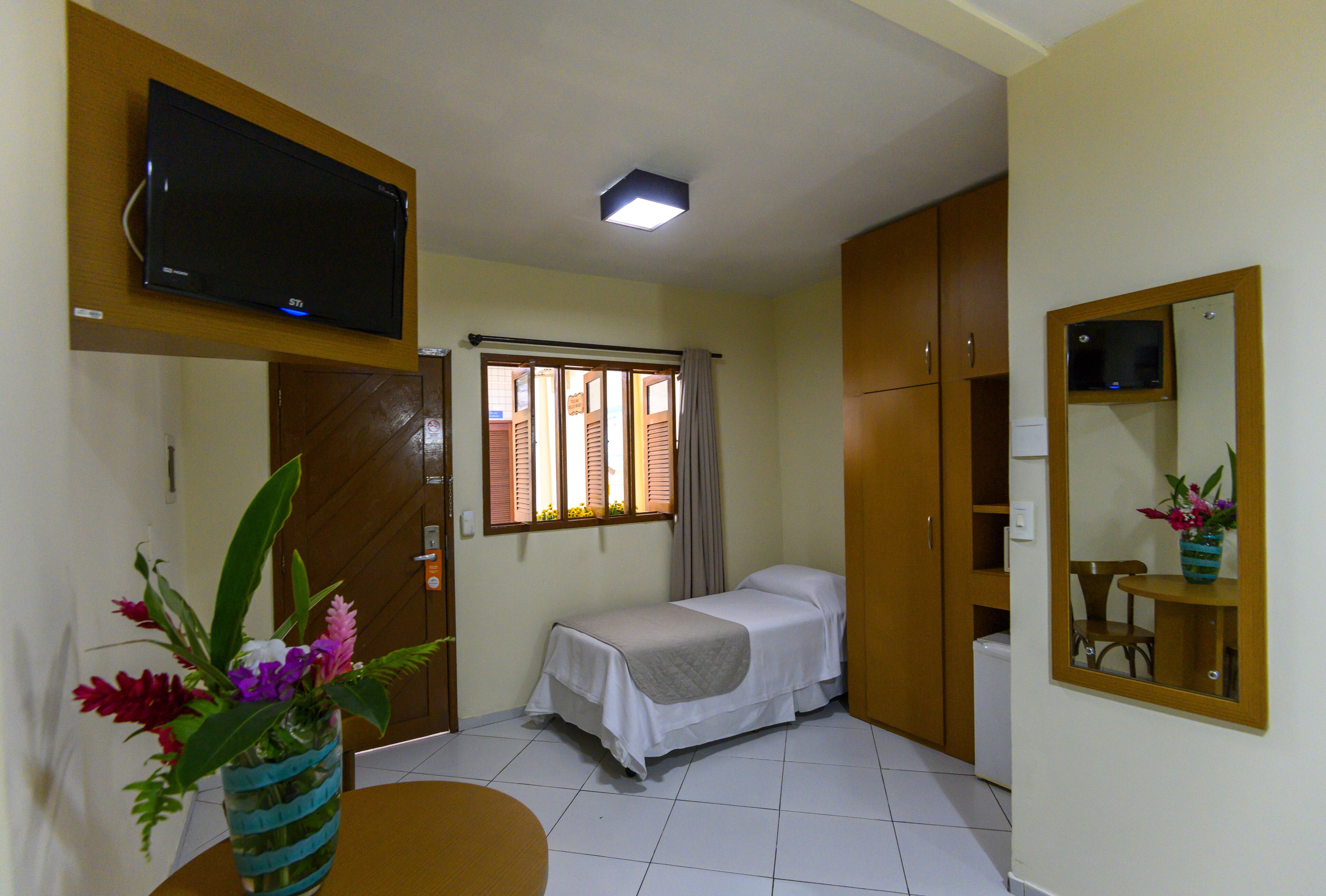 A small, clean room with a bed, a TV mounted on the wall, a window with curtains, and a mirror on the wall.