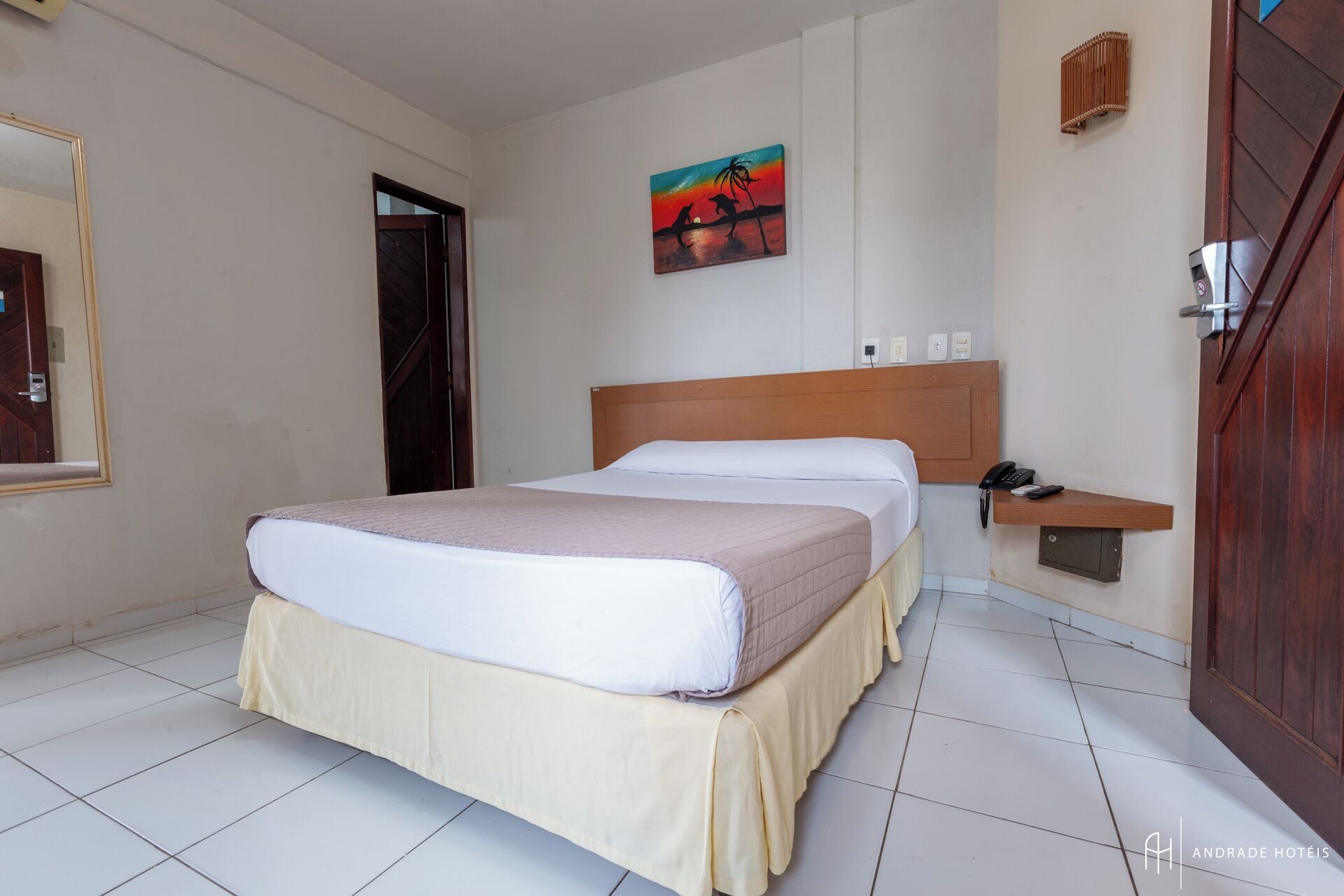 A hotel room with a bed, a painting, a phone, and a small shelf.