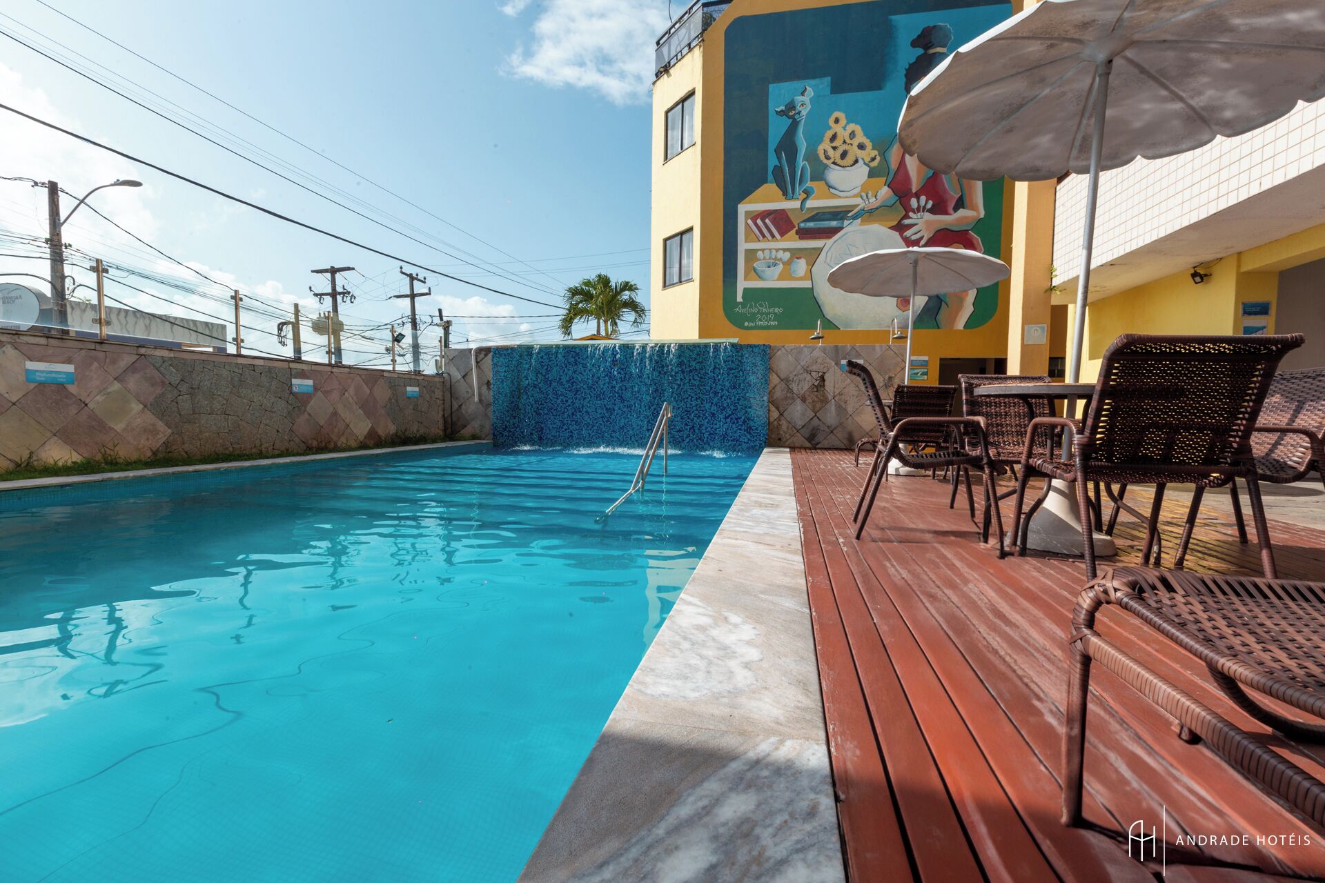 Una piscina con borda piastrellata, circondata da sedie e tavoli in vimini, e un murale sull'edificio adiacente.