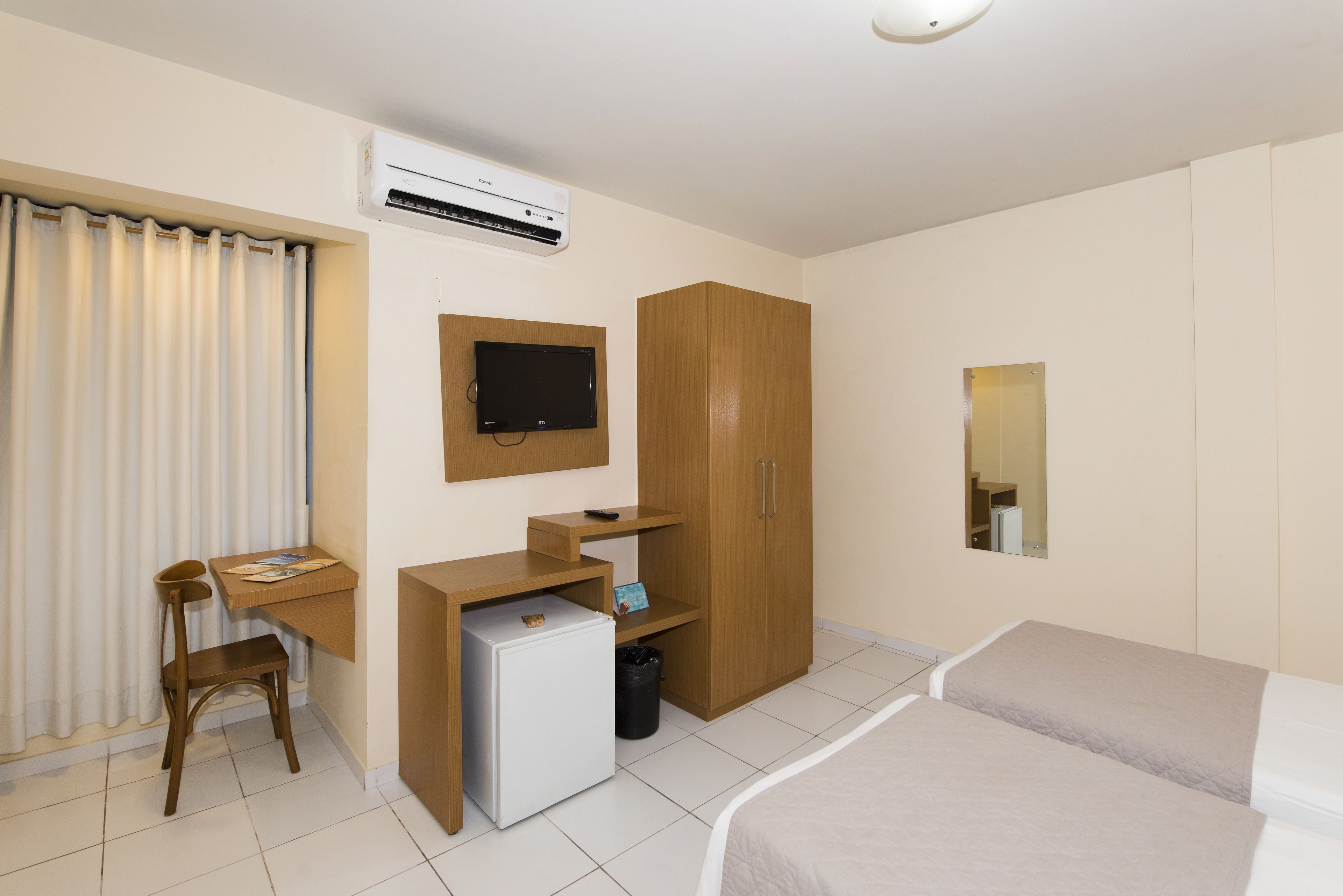 Quarto de hotel com cama, secretária com computador, pequeno frigorífico, televisão na parede e guarda-roupa.