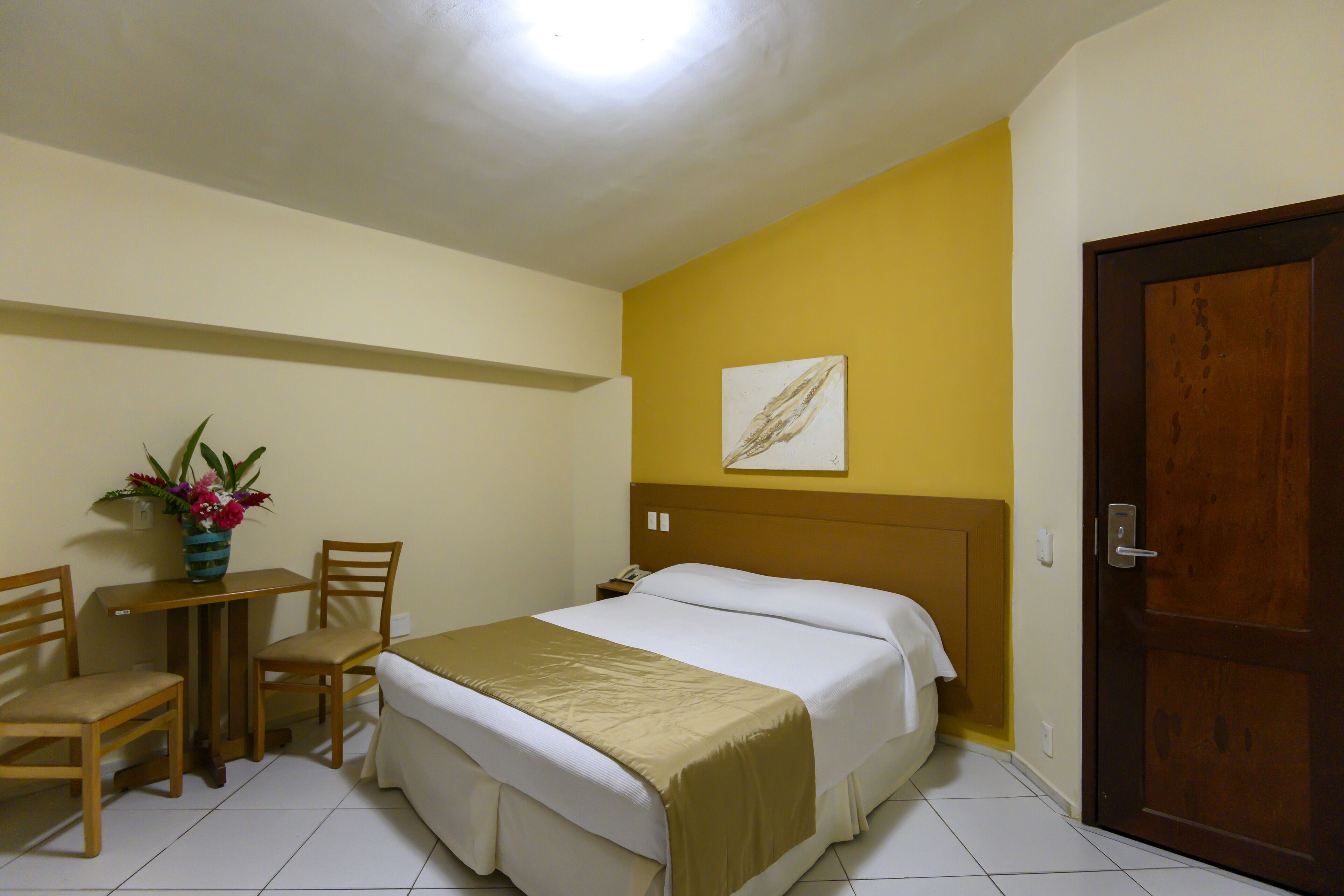 Una camera d'albergo con un letto, una scrivania, due sedie, un tavolino con un vaso di fiori e una porta.