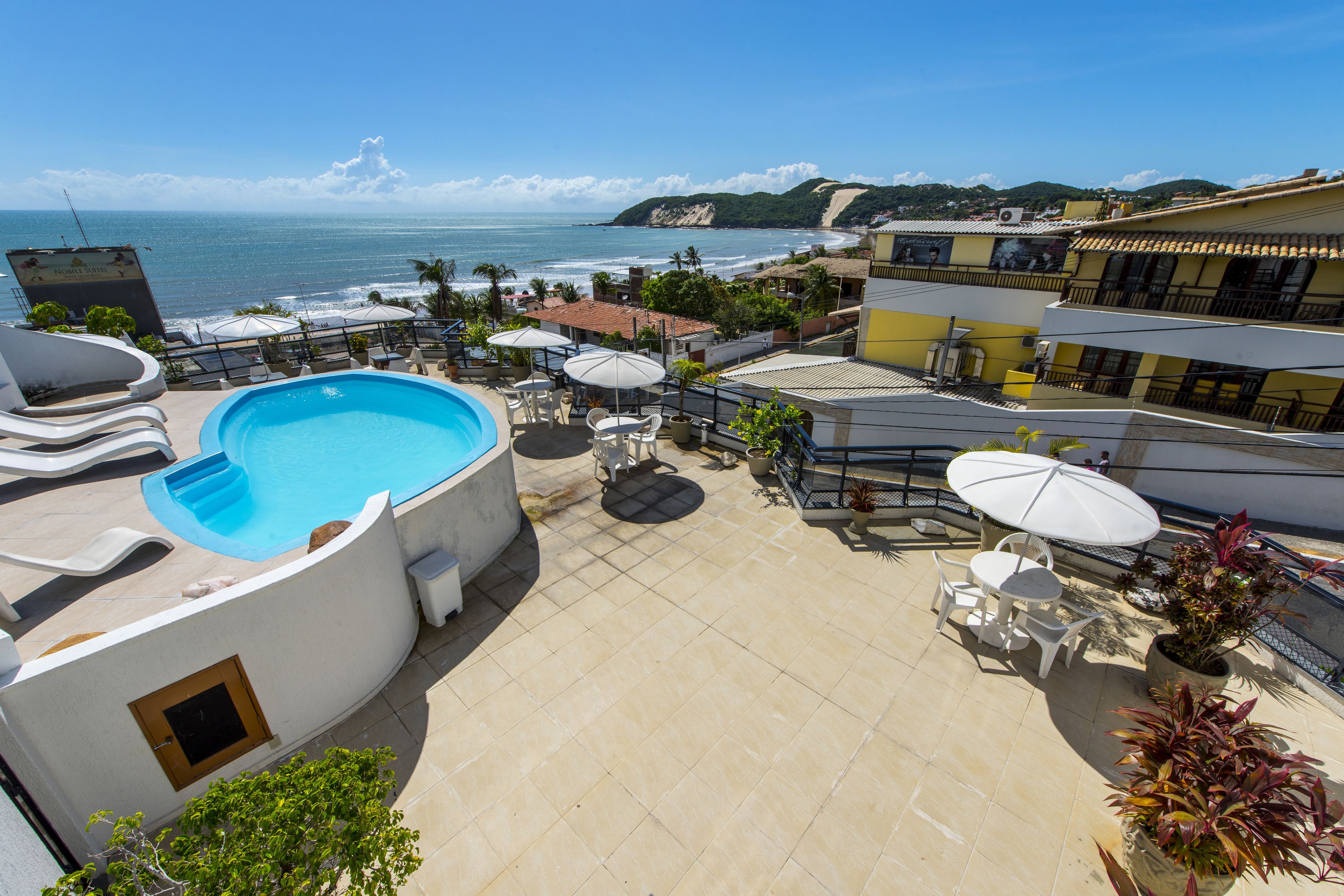 A rooftop pool area with a view of the ocean, outdoor seating, and a building with a yellow facade.