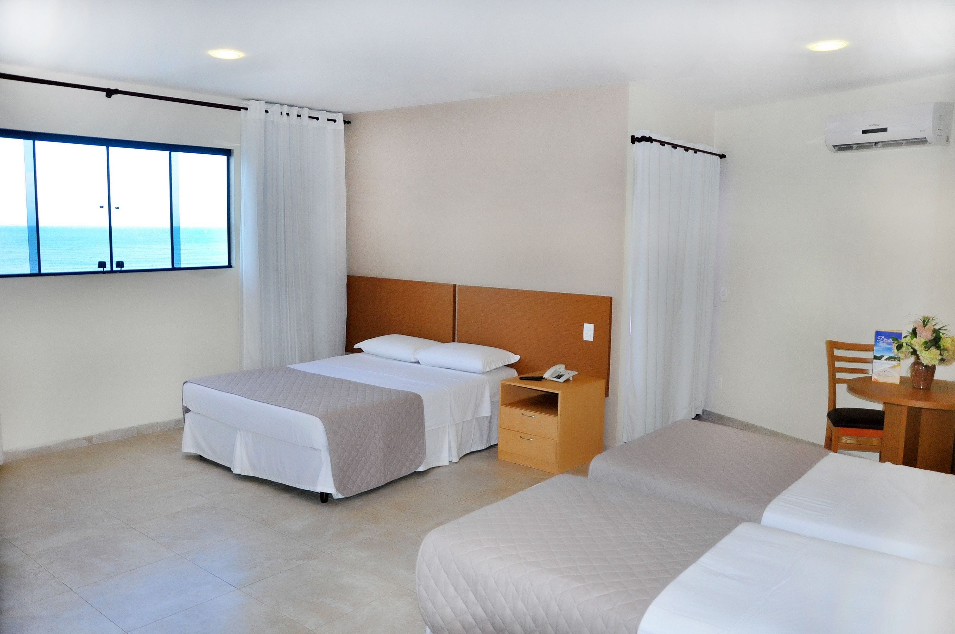 A hotel room with a large window, a bed, a bedside table with a phone, and a chair with a table.