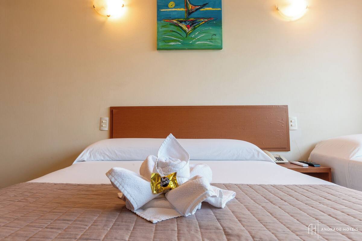 Quarto de hotel com cama bem arrumada, um quadro na parede e mesa de cabeceira com telefone e um livro.