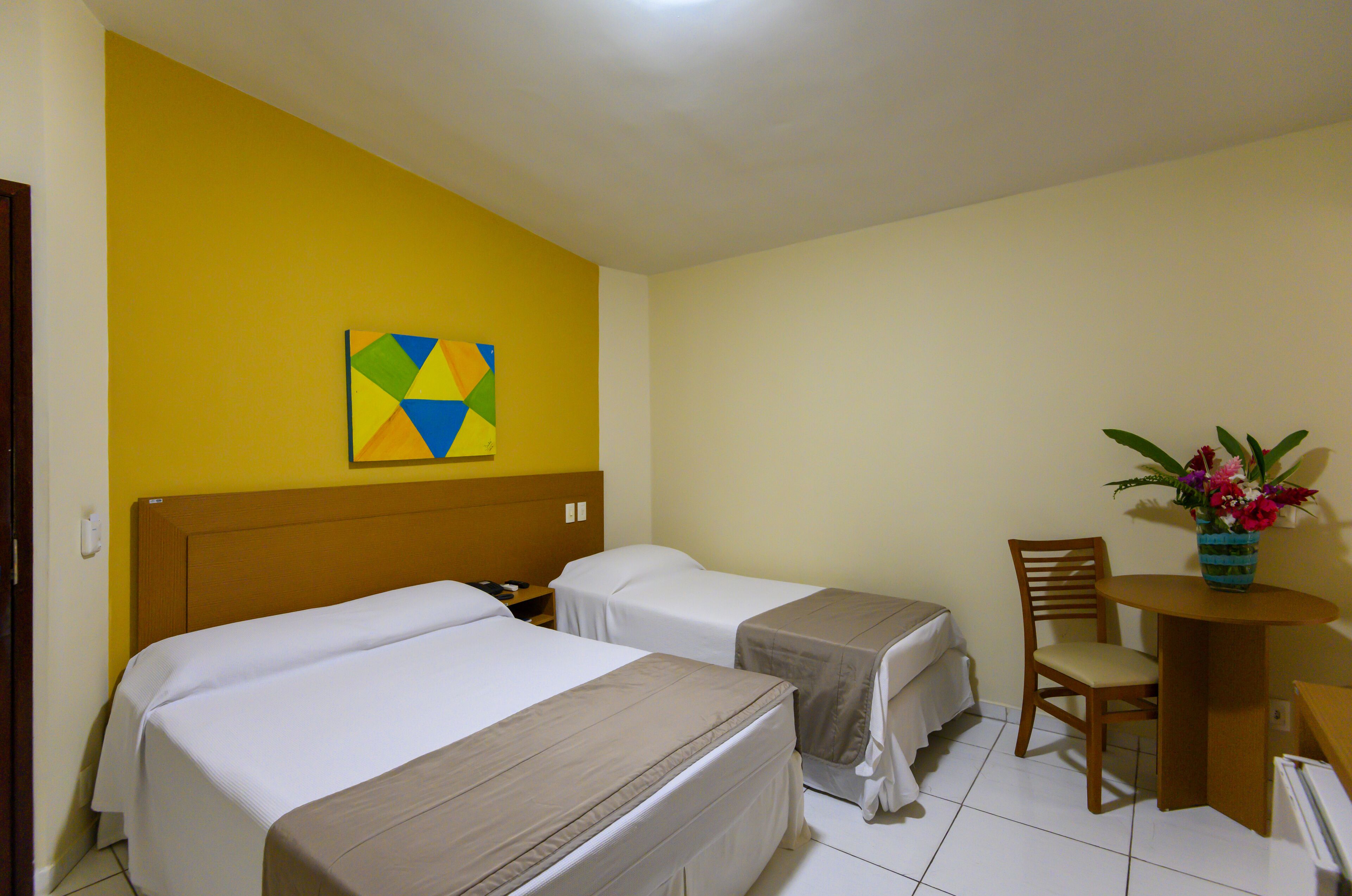 A hotel room with two beds, a wooden headboard, a small table with a chair, and a colorful painting on the wall.
