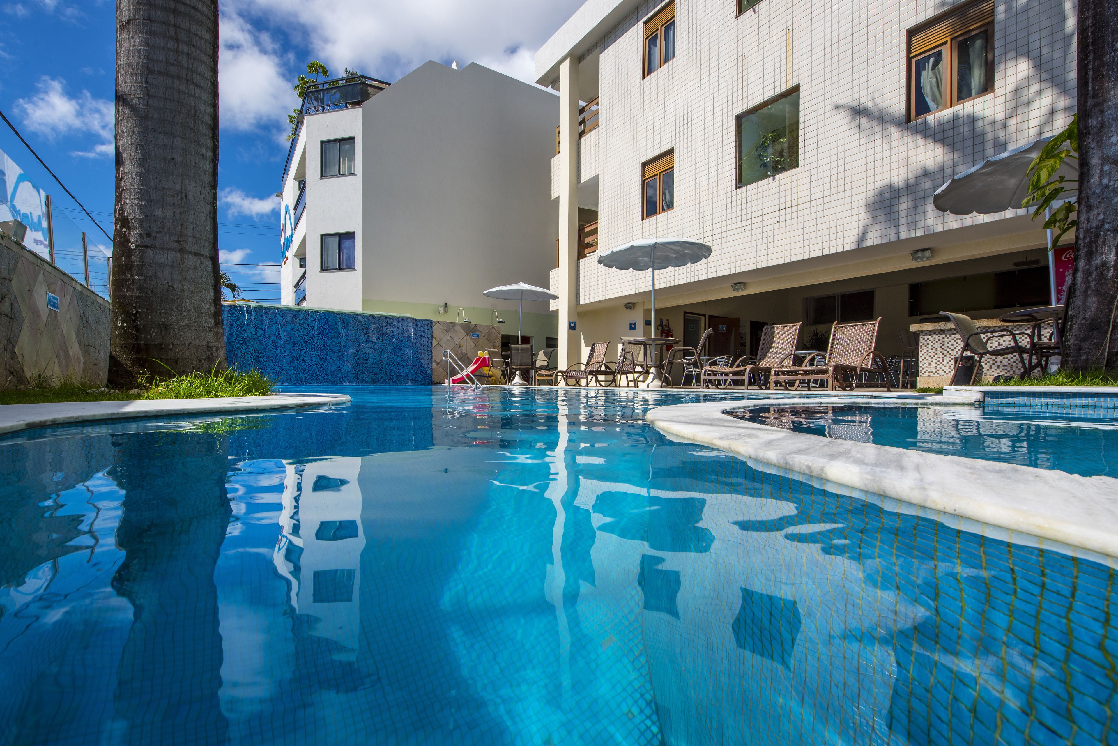 A swimming pool with lounge chairs and umbrellas, surrounded by a building with tiled walls and balconies.