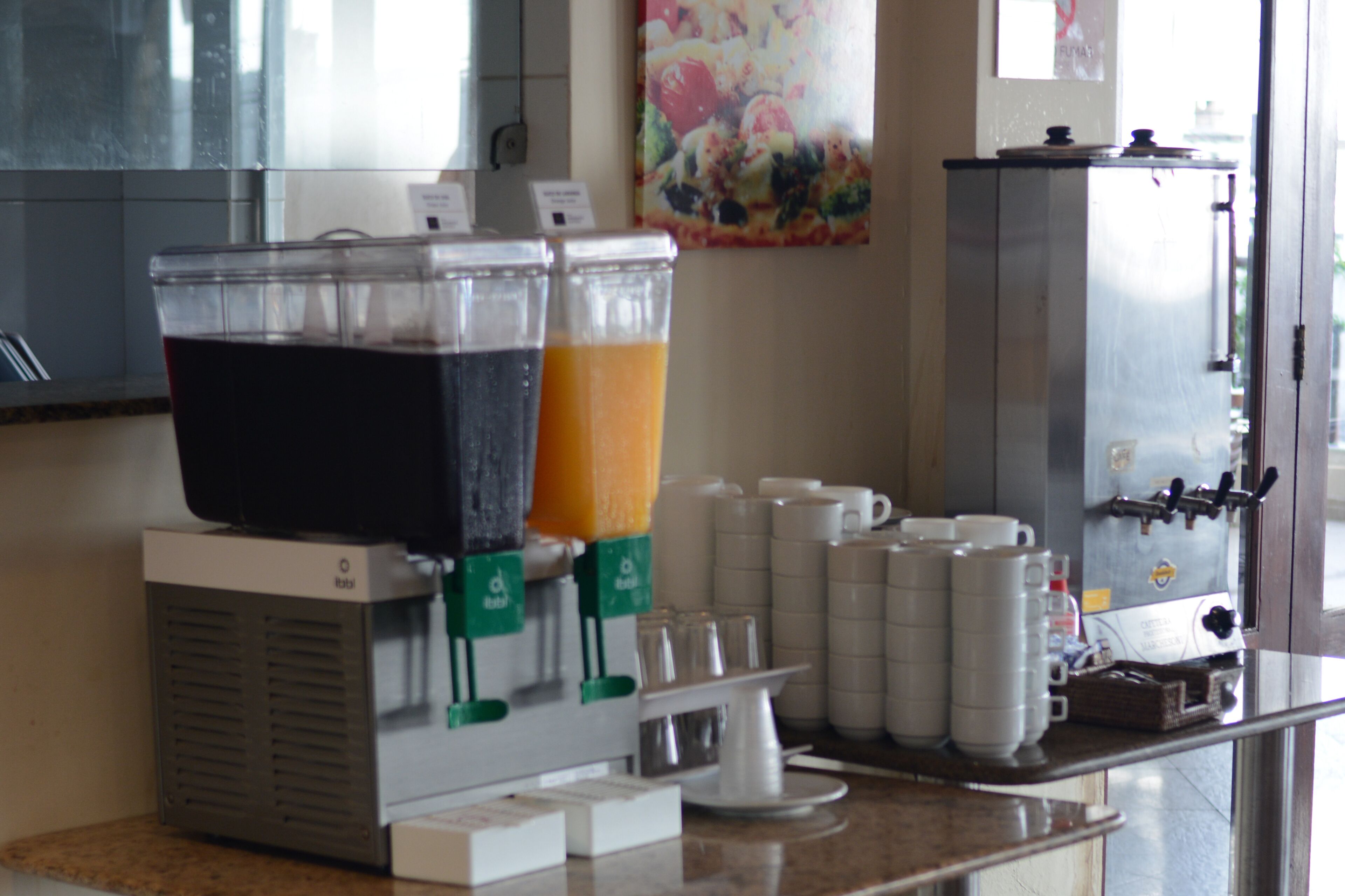 Una estación de bebidas con varios dispensadores, una máquina de café y vasos apilados.