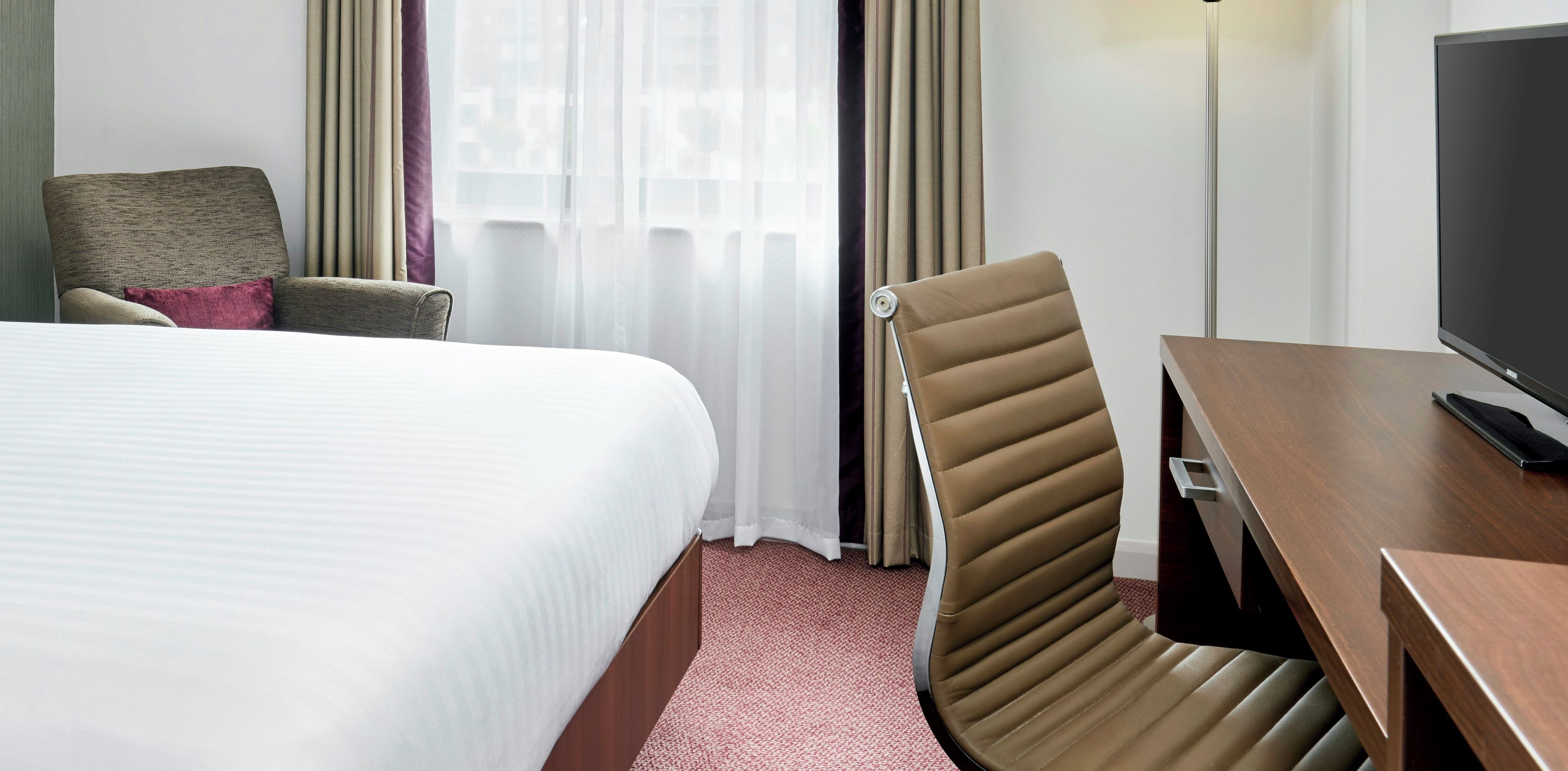 A hotel room with a bed, a desk with a television, a chair, and a window with curtains.