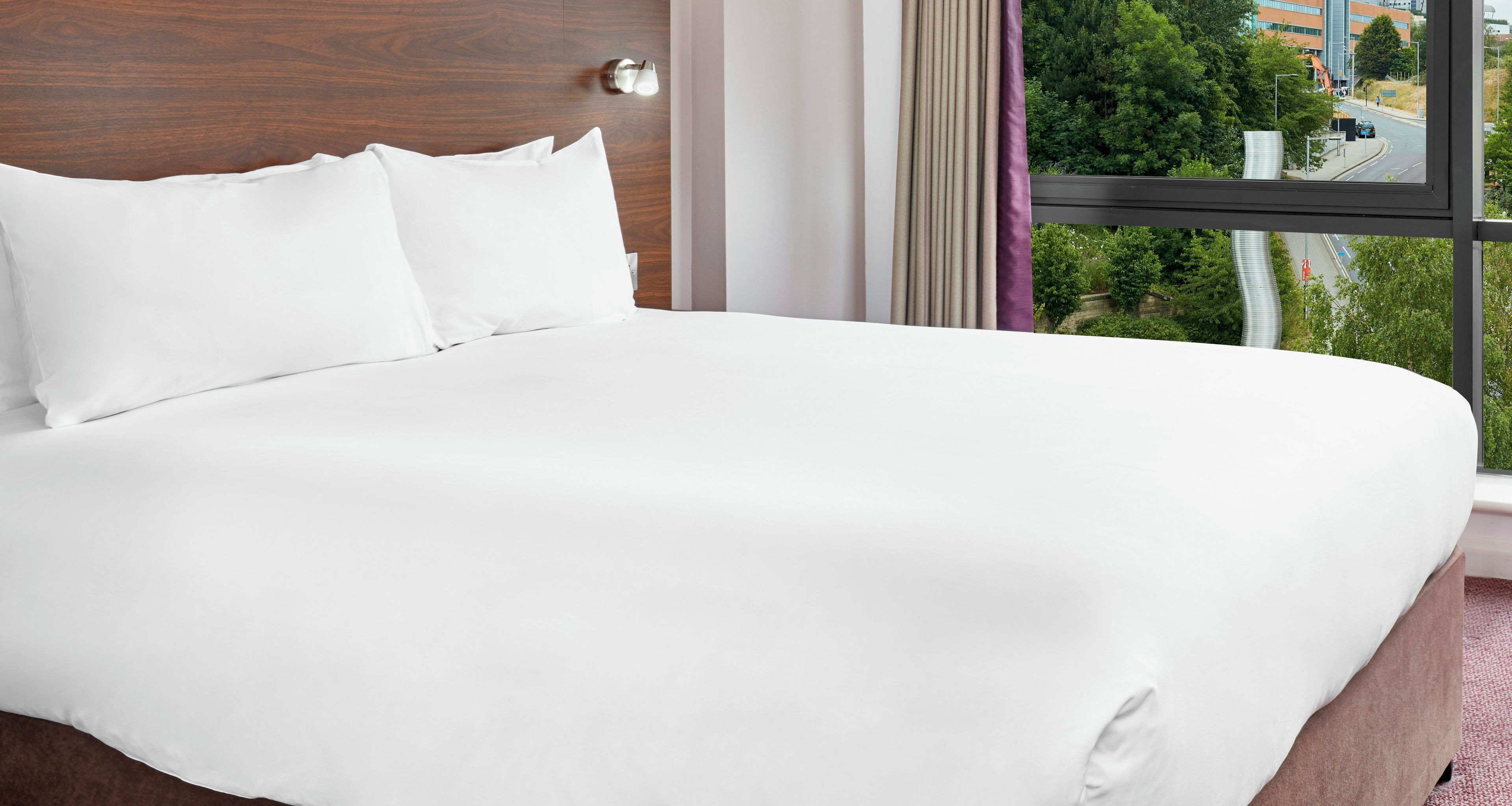 Una cama bien hecha con ropa blanca en una habitación de hotel con un ventanal grande.