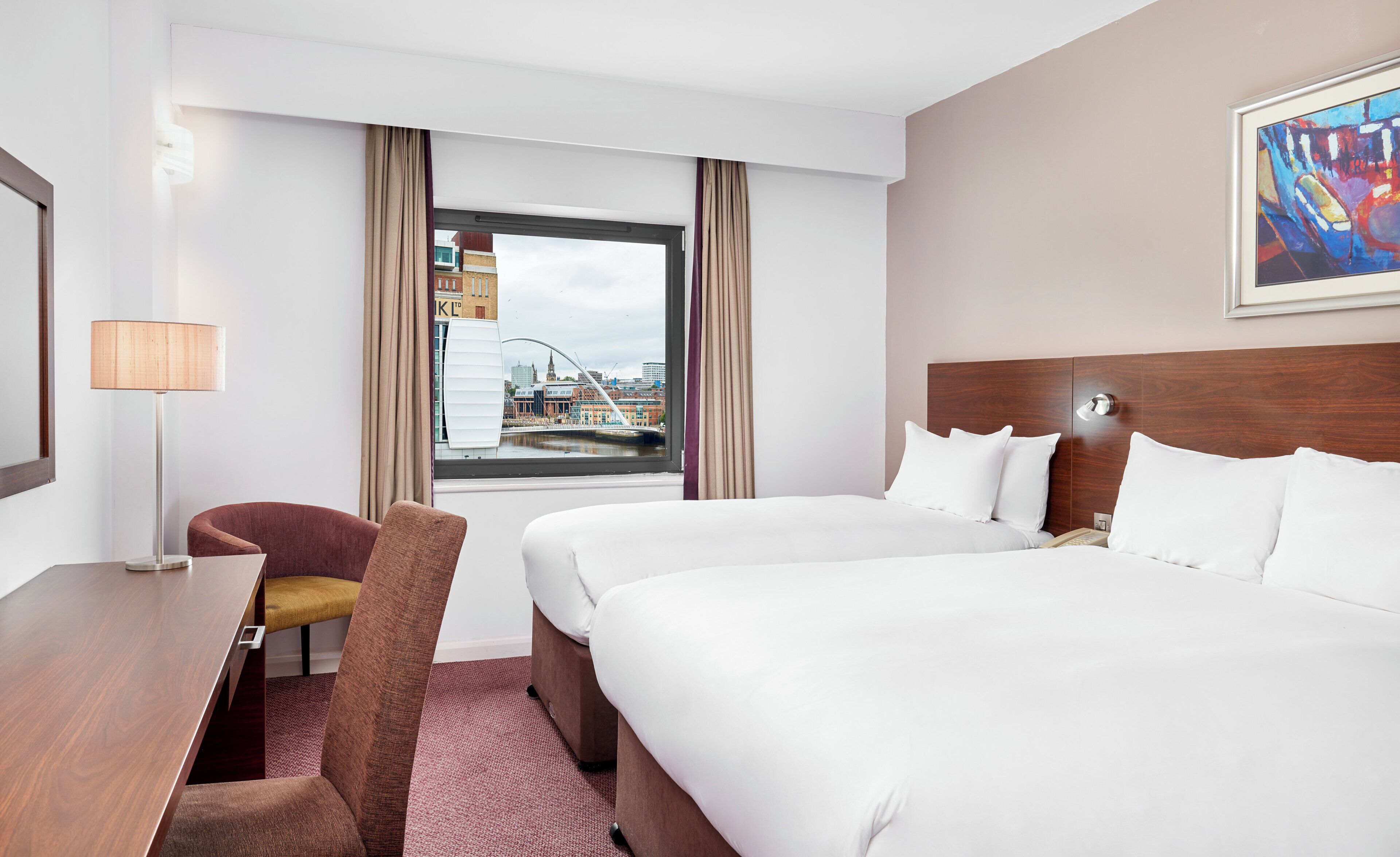 Une chambre d’hôtel avec un grand lit, un bureau avec une lampe, une chaise et une vue sur le paysage urbain par la fenêtre.