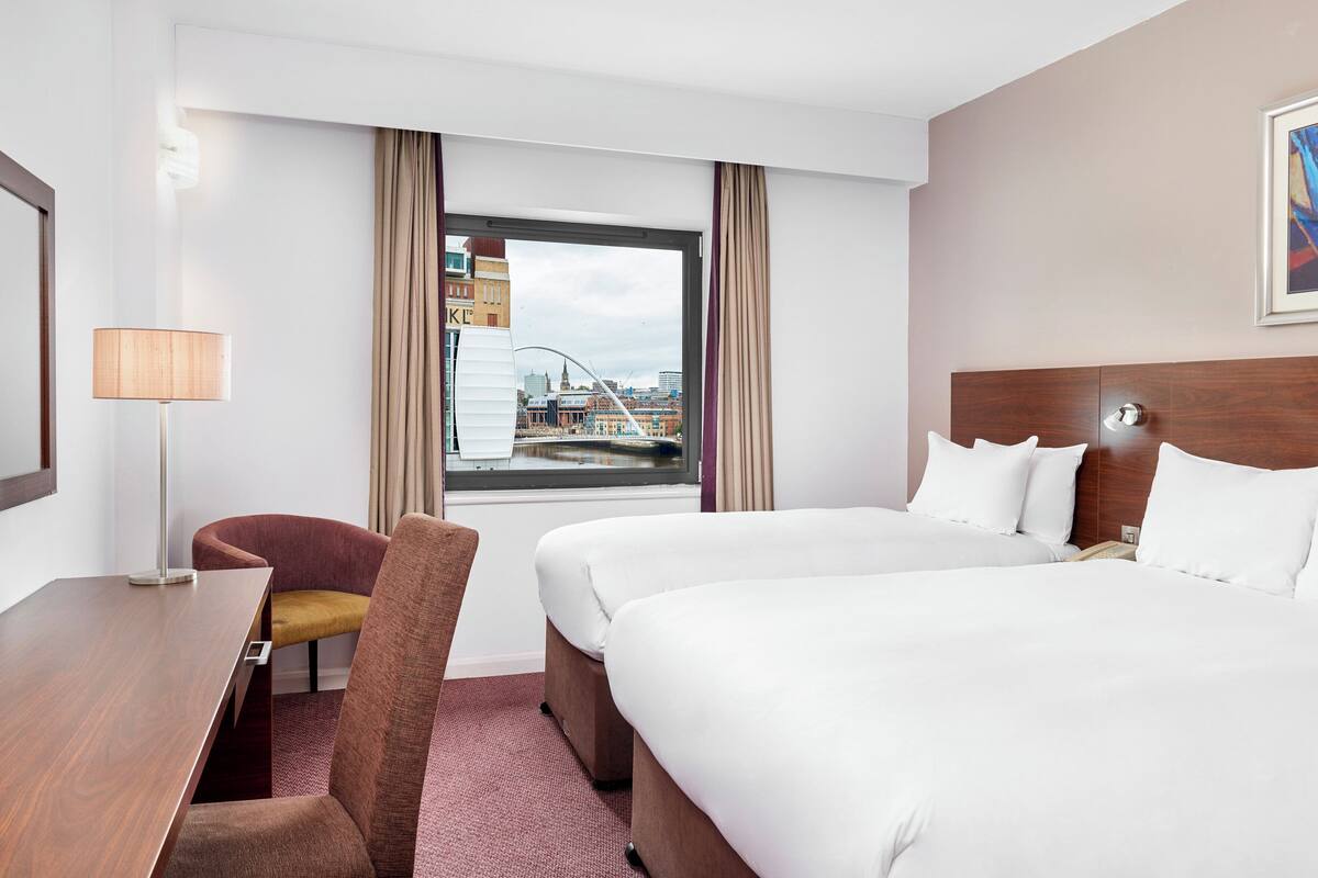 Un cuarto de hotel con una cama grande, un escritorio con una lámpara, una silla y vistas de un paisaje urbano a través de la ventana.