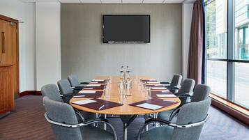 Un salón de conferencias con una gran mesa, sillas grises, un televisor de pantalla plana y vistas al exterior a través de amplios ventanales.