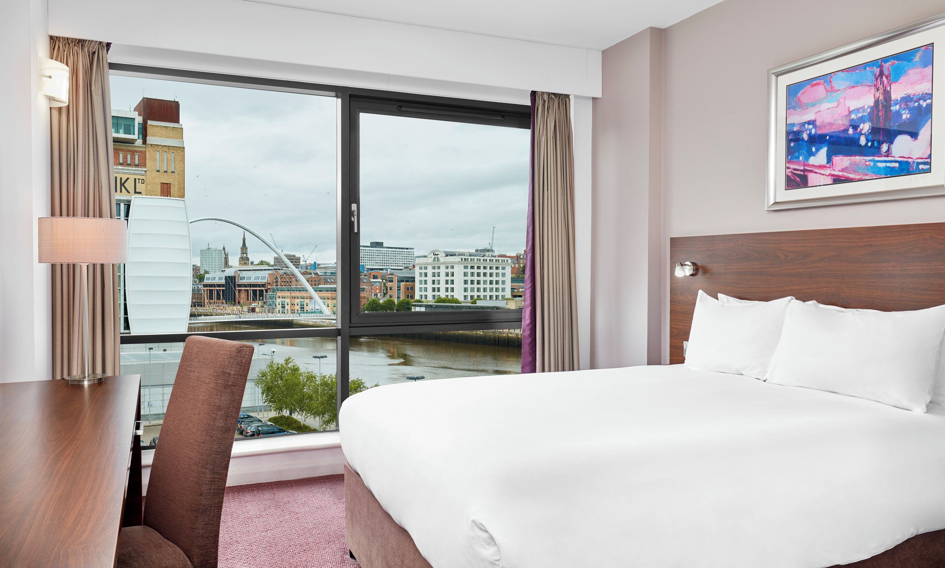Habitación de hotel con una cama grande, un escritorio, una silla y vistas a un paisaje urbano a través de un ventanal amplio.