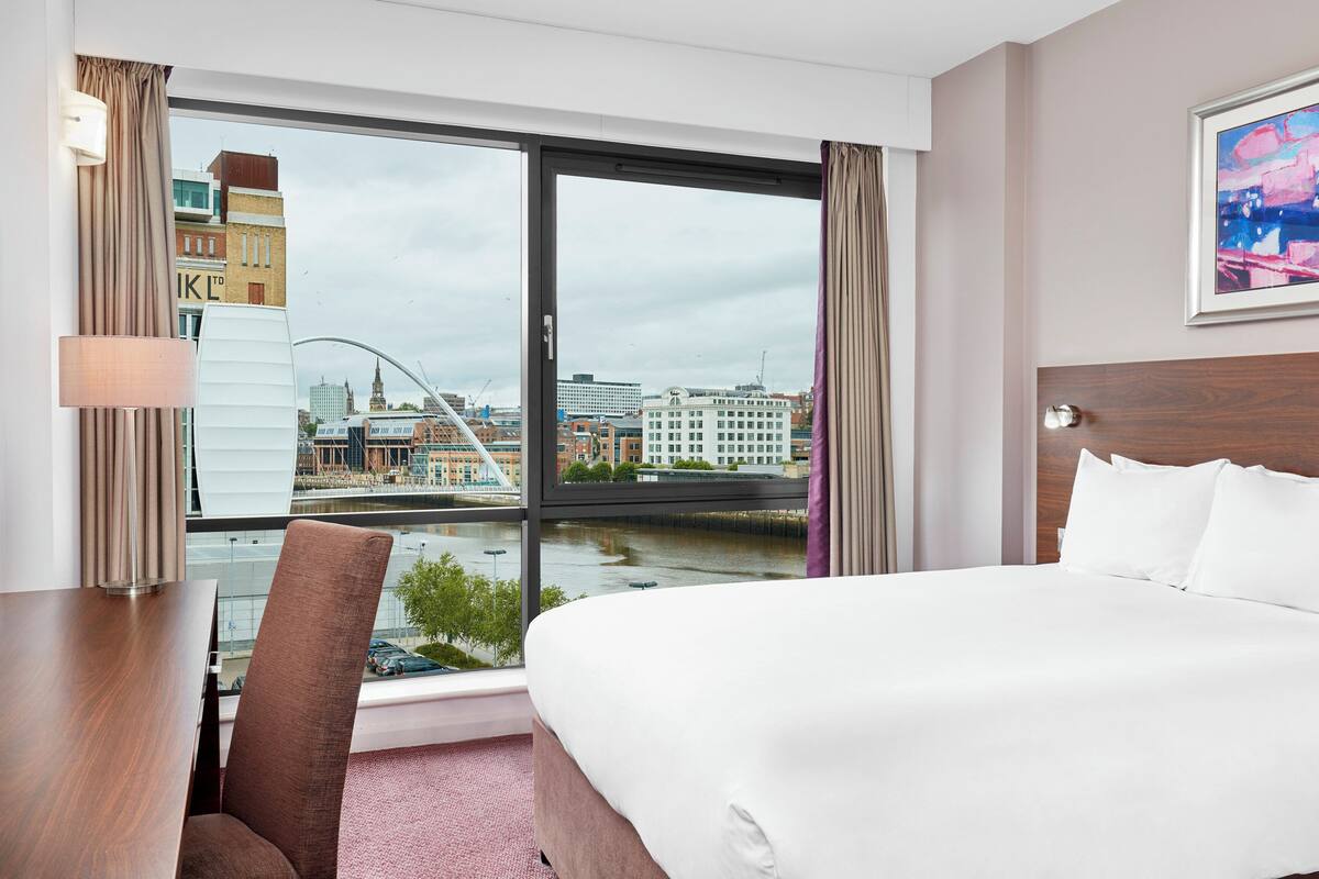 Habitación de hotel con una cama grande, un escritorio, una silla y vistas a un paisaje urbano a través de un ventanal amplio.