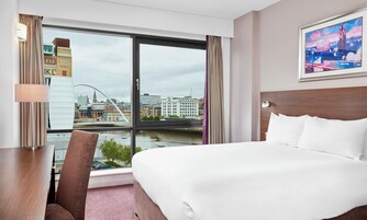 Habitación de hotel con una cama grande, un escritorio, una silla y vistas a un paisaje urbano a través de un ventanal amplio.