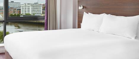 Habitación de hotel con una cama grande, un cabecero de madera y vistas a un paisaje urbano a través de una ventana.