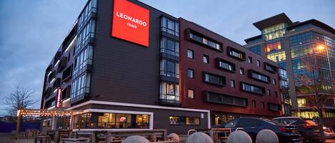 A modern hotel with a prominent 'Leonardo Hotels' sign, outdoor seating, and a parking area.