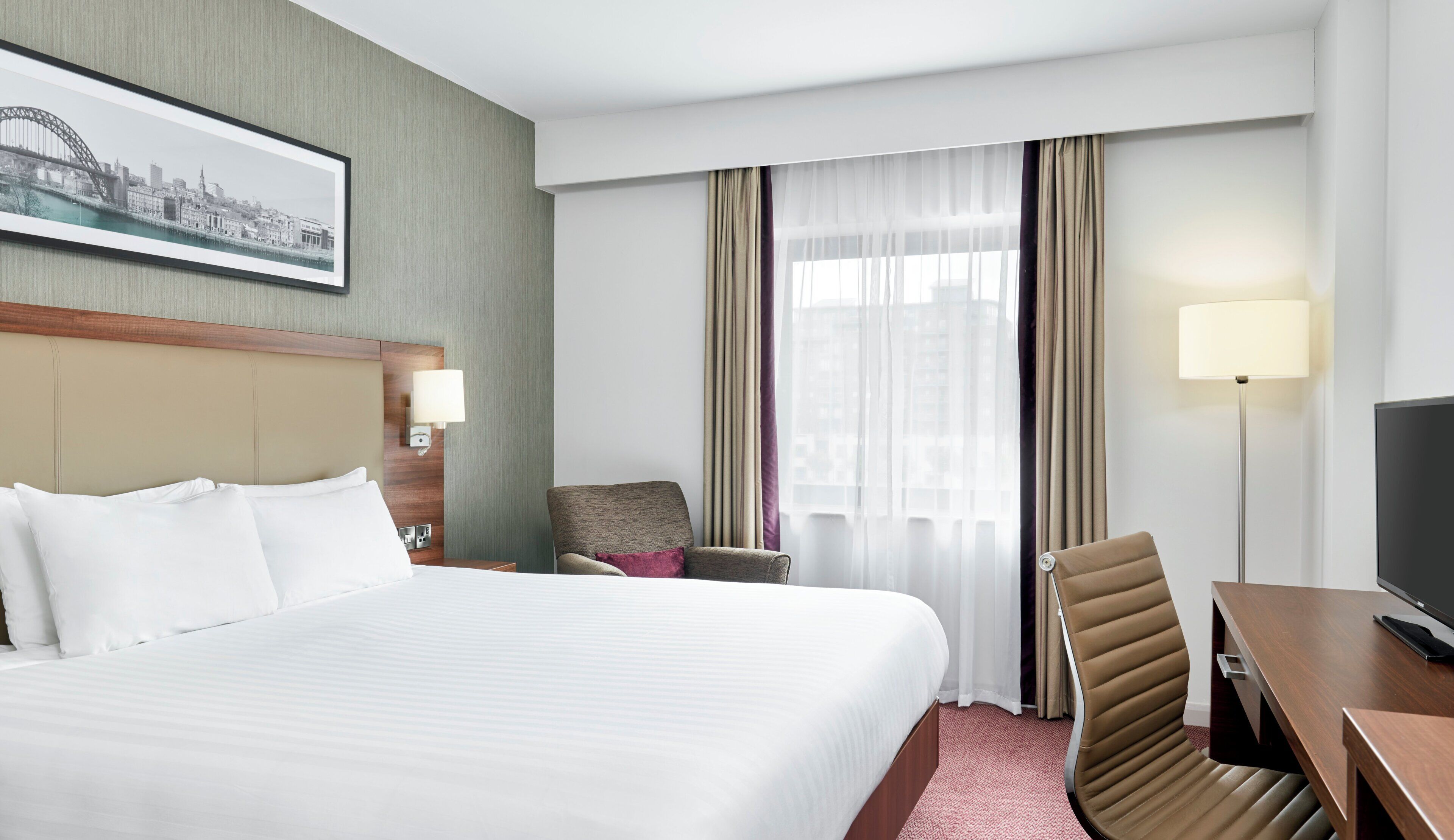 A hotel room with a large bed, a desk with a television, a chair, a nightstand with a lamp, and a window with curtains.