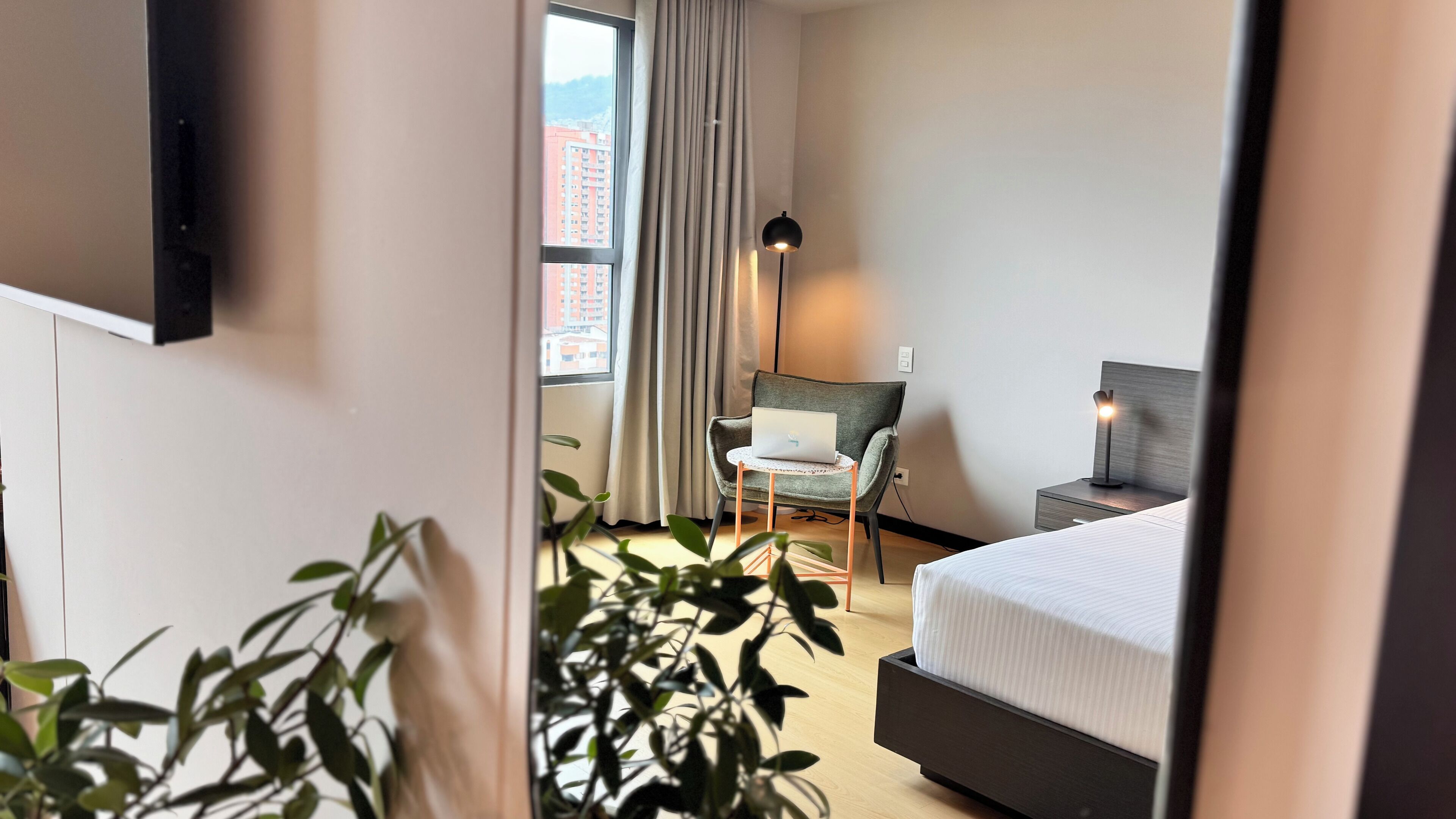 Ein Hotelzimmer mit Bett, Schreibtisch mit Laptop, Stuhl, Stehlampe und Blick auf Gebäude durch das Fenster.