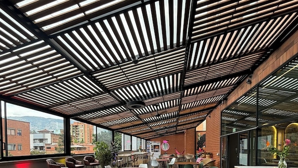 Un espace de restauration intérieur doté de nombreuses tables et chaises, d’un plafond haut avec des lambris en bois et de grandes fenêtres offrant une vue sur les bâtiments environnants.