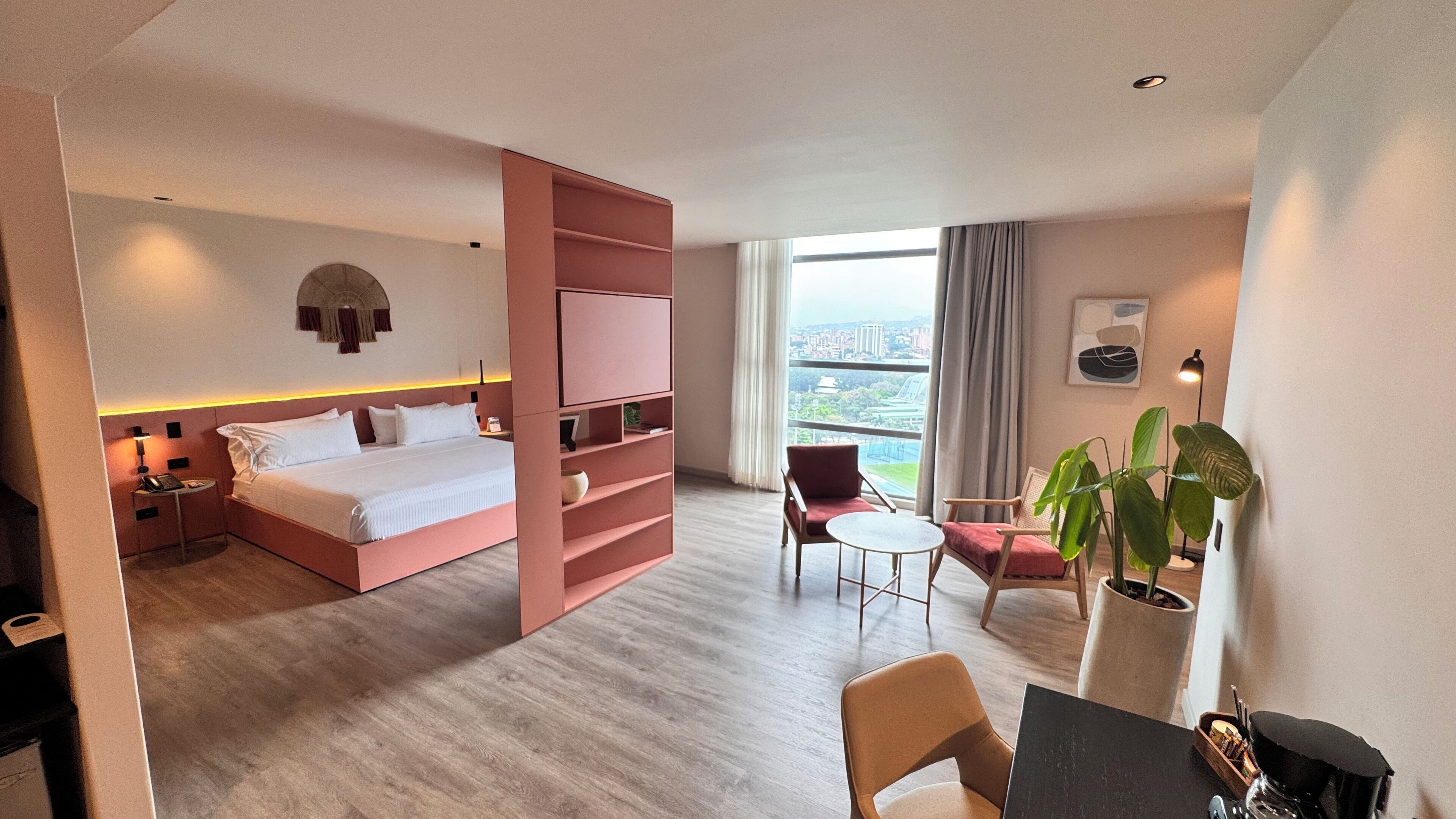 A modern hotel room with a large bed, a desk, a chair, a TV, and a view of the city.