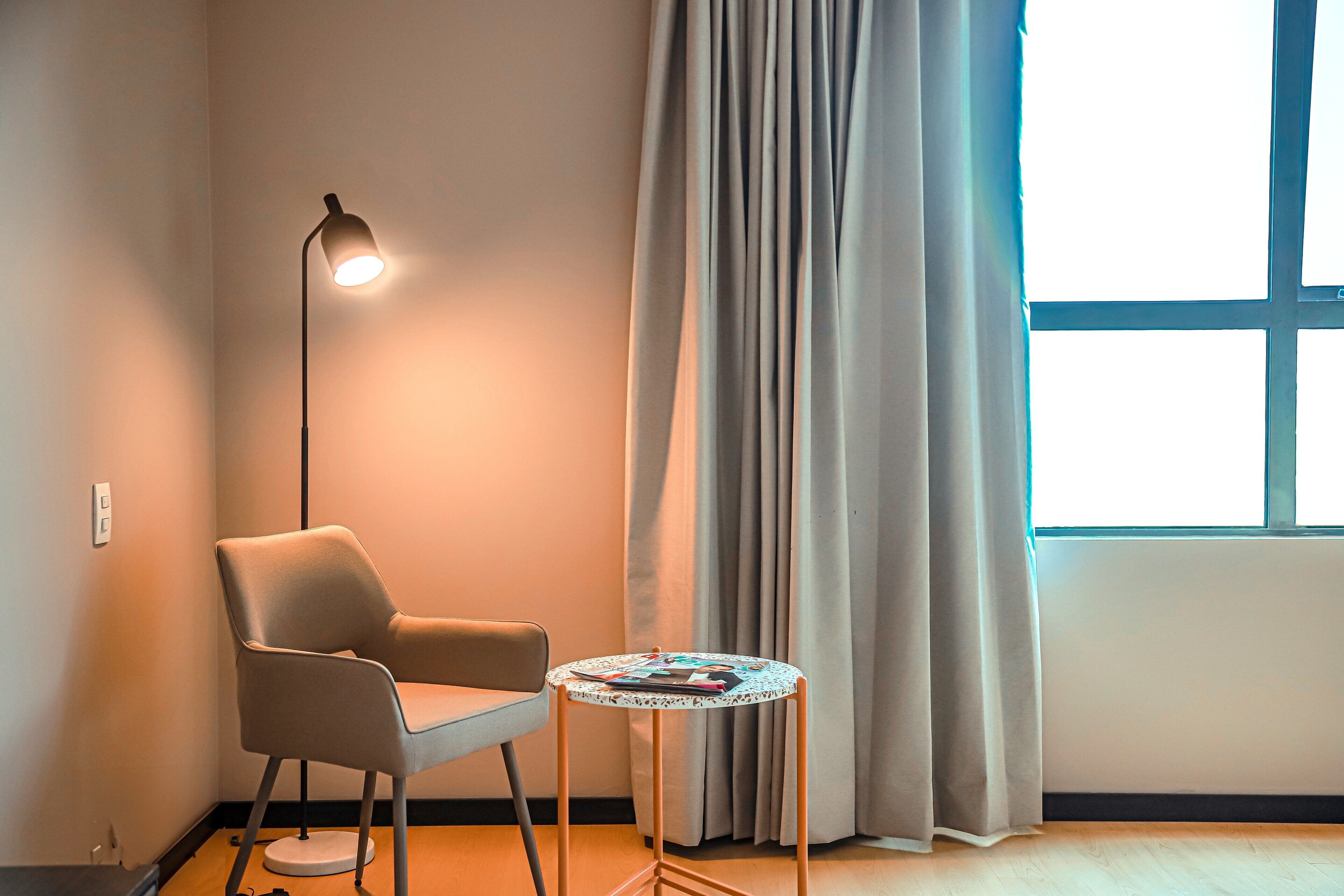 Quarto com uma cadeira, uma pequena mesa, um abajur e uma janela com cortinas.