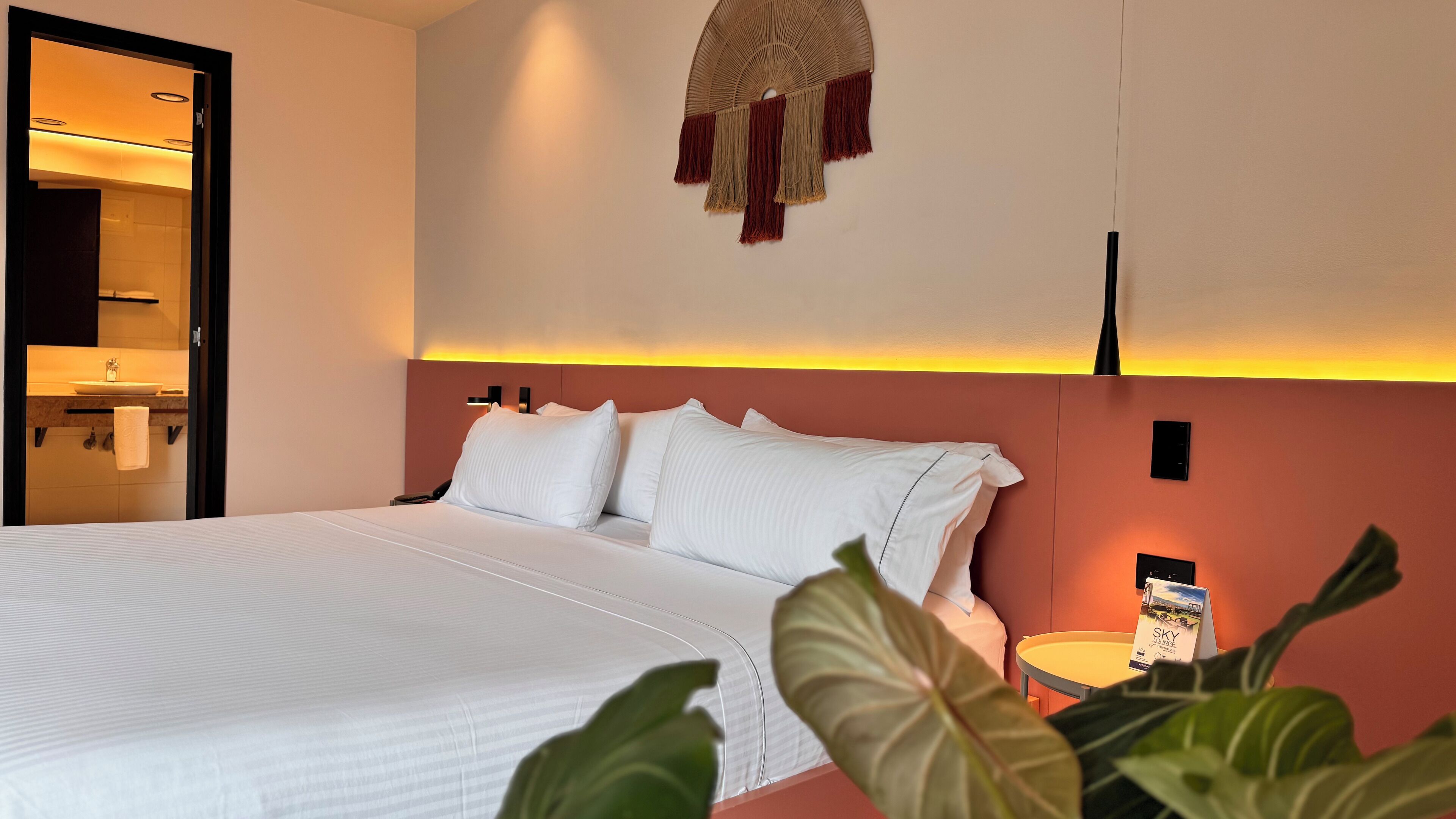 Et hotelværelse med en stor seng, et sengebord med lampe, en vægmonteret ventilator og udsigt til badeværelset.