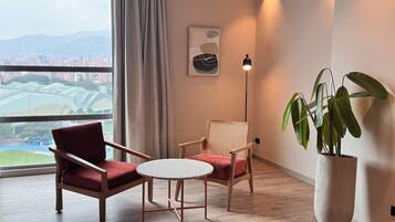 A room with two chairs, a small round table, a potted plant, a floor lamp, and a city view through a large window.