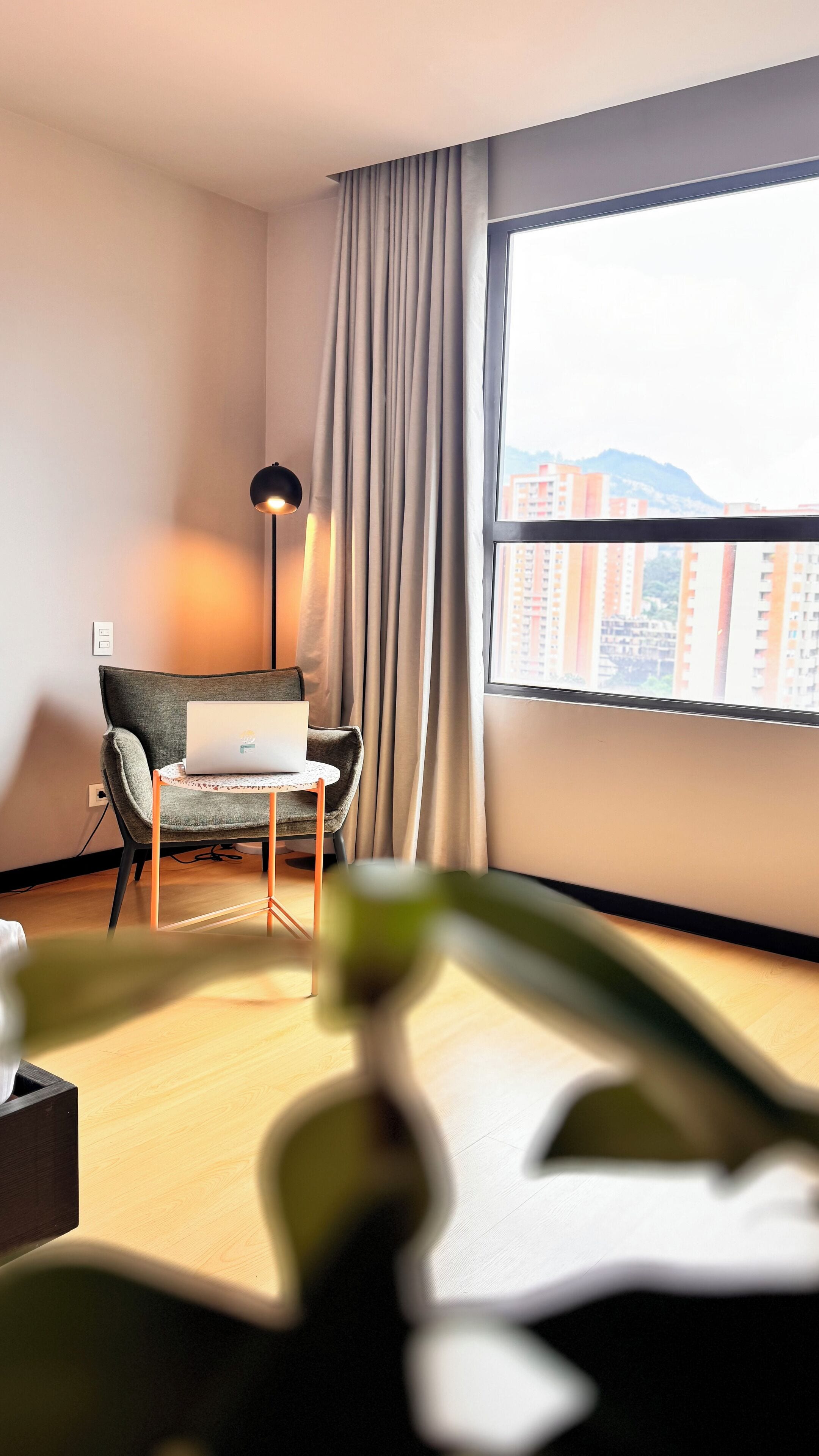 Quarto de hotel moderno com vista para o horizonte urbano através de uma grande janela, uma poltrona cinza, uma pequena mesa redonda com um laptop e um abajur.