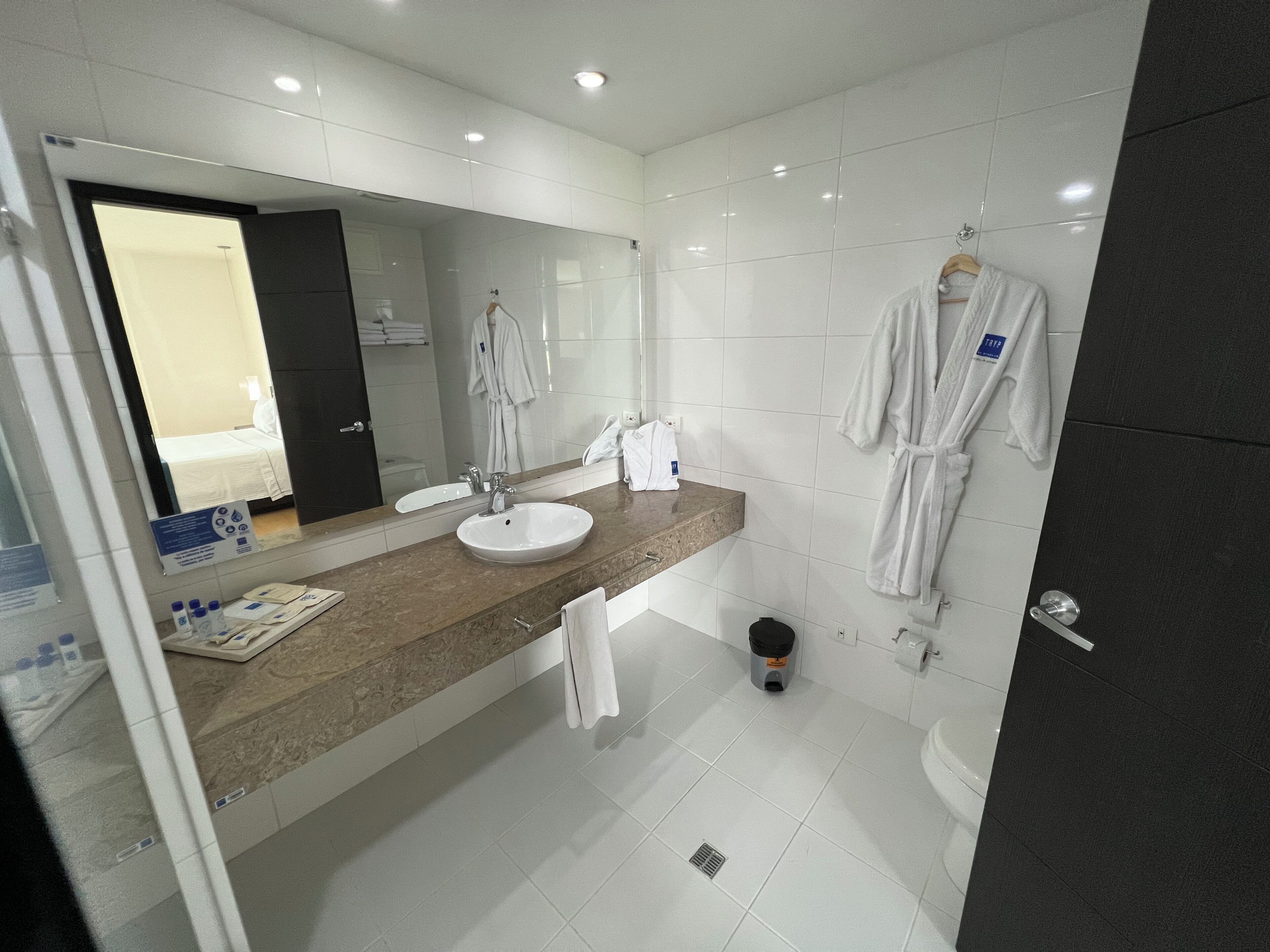 Une salle de bain moderne avec un grand miroir, un lavabo et des toilettes. Des serviettes blanches et un peignoir sont accrochés au mur.