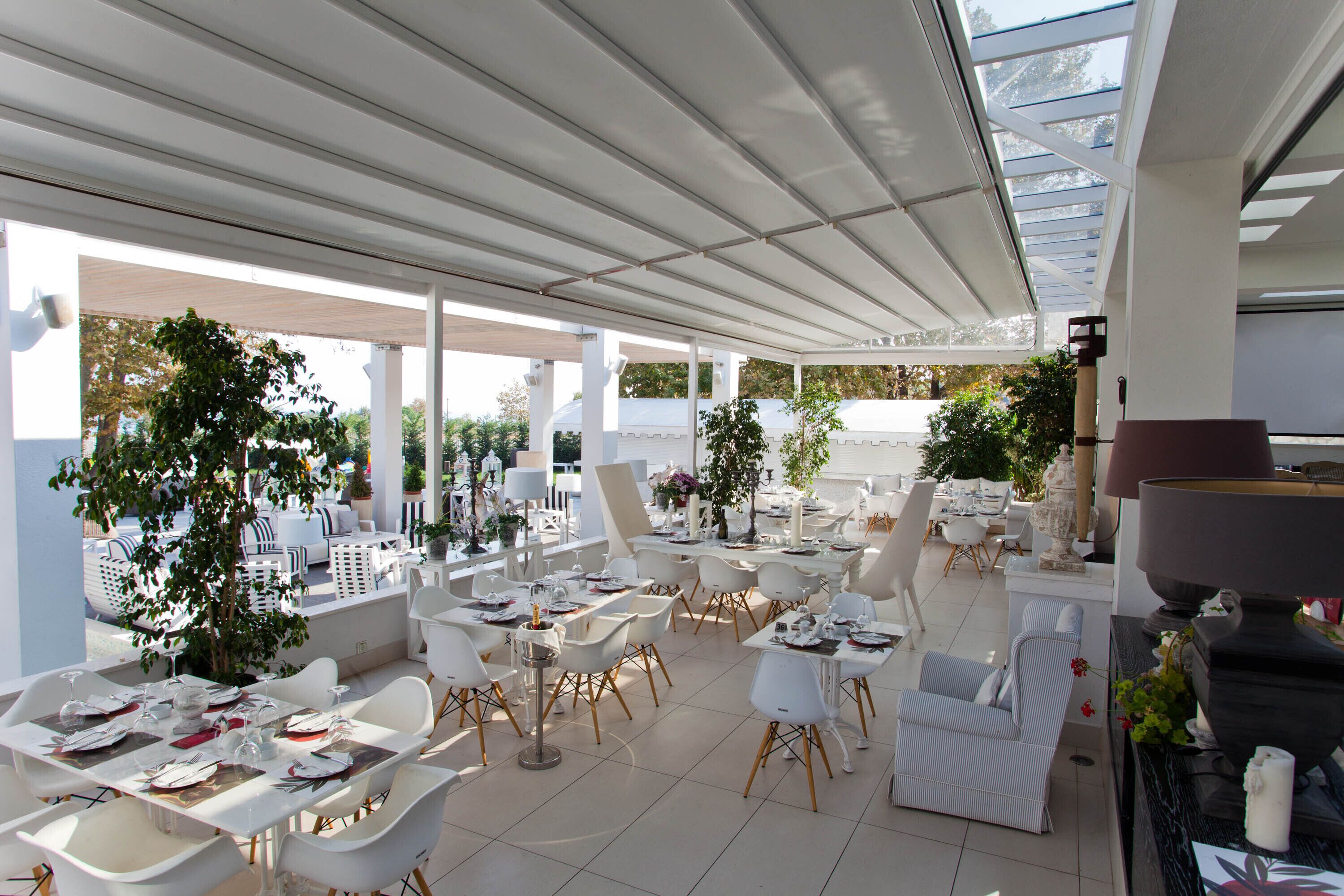 An outdoor dining area with white tables and chairs, surrounded by potted plants and a covered patio.