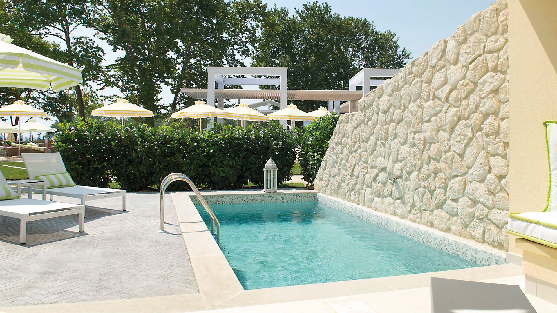 A pool area with lounge chairs, umbrellas, and a stone wall.