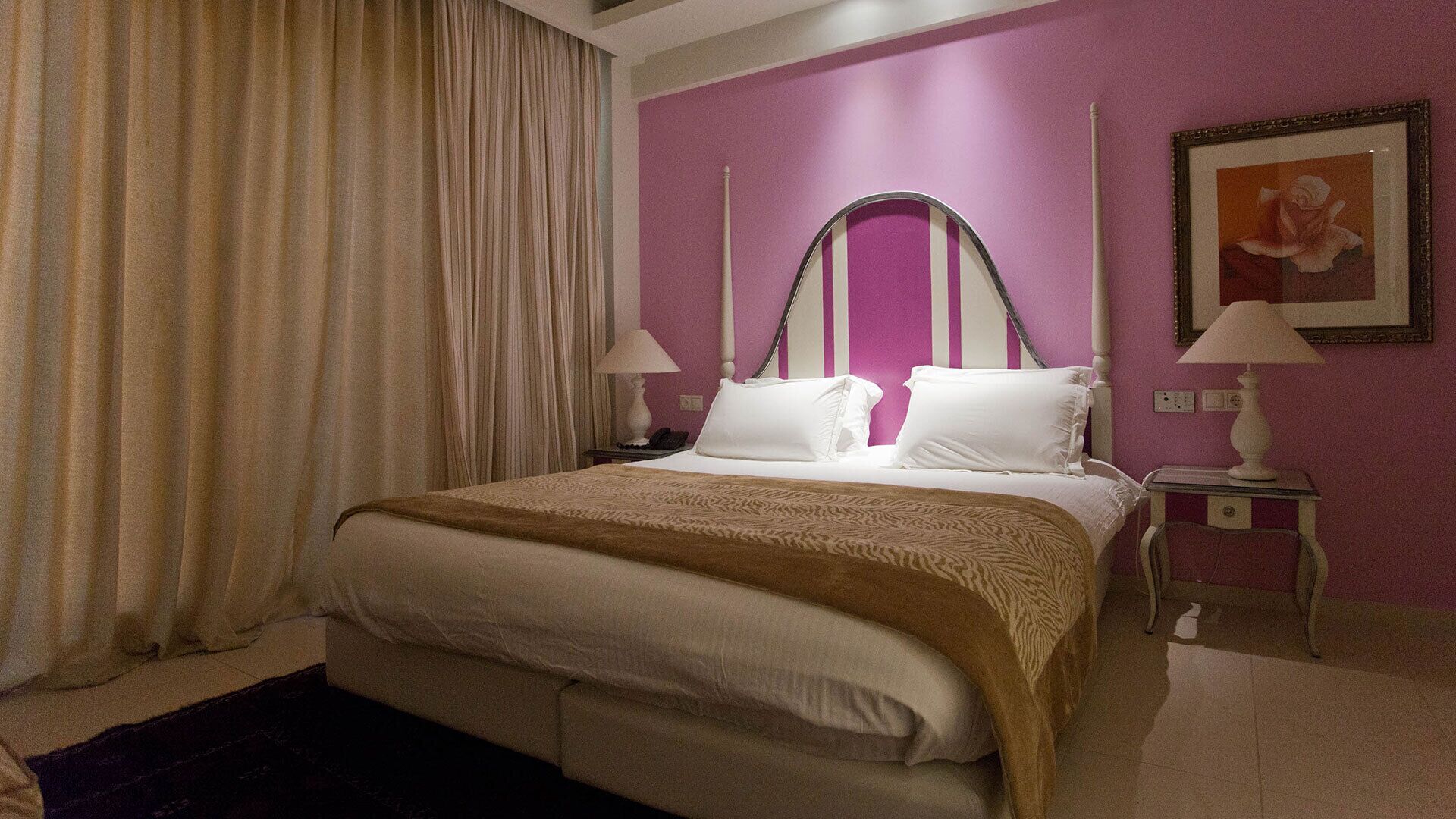 Habitación de hotel con cama, mesita de noche, lámpara, un cuadro en la pared y ventana con cortinas.