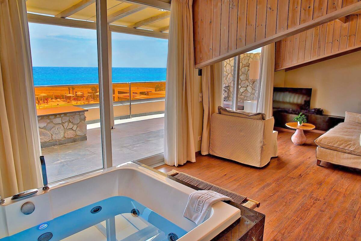 Ein modernes Wohnzimmer mit großem Fenster mit Blick auf einen Strand, einer Whirlwanne und einer gemütlichen Sitzecke.