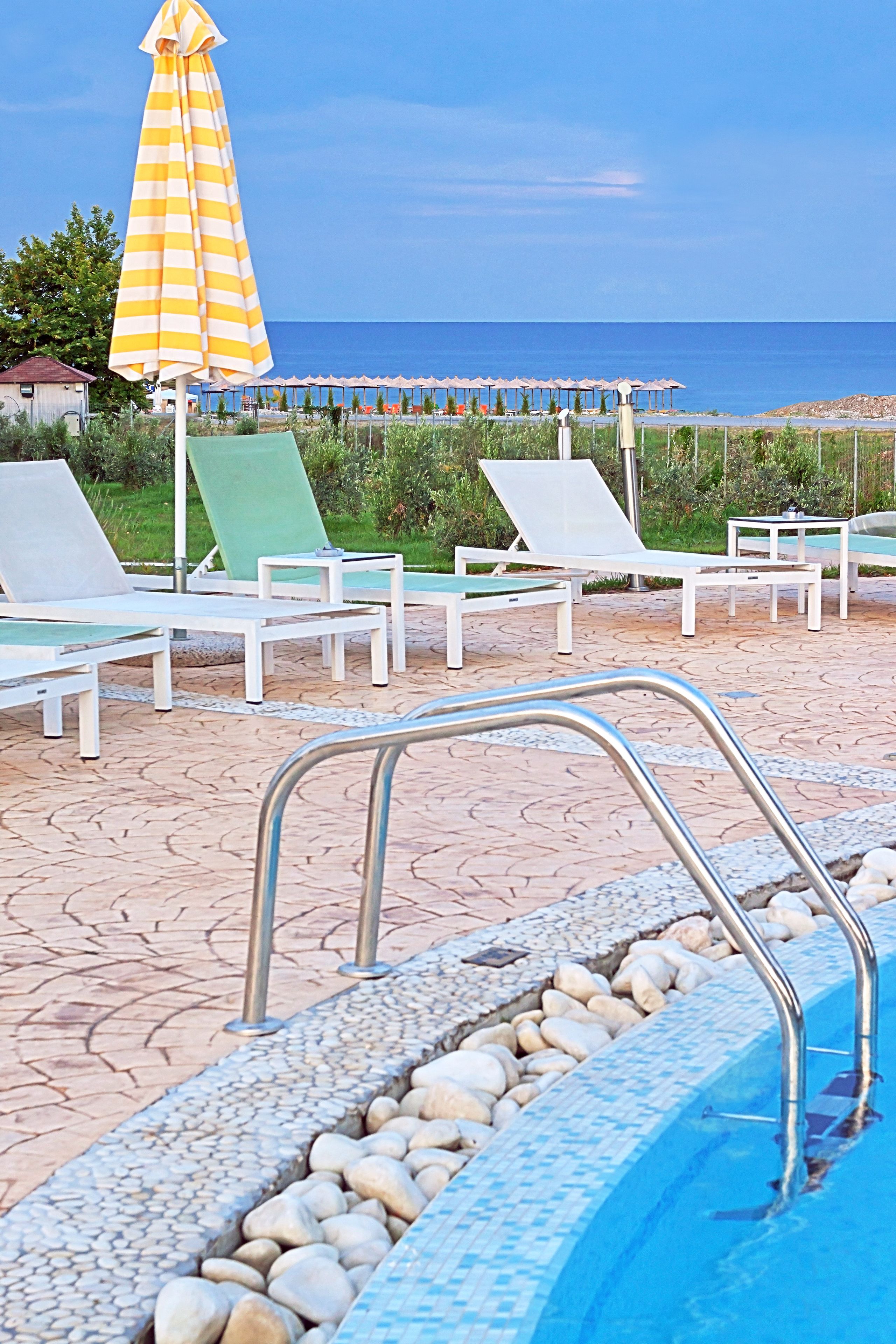 A pool area with lounge chairs and a striped umbrella, a clear view of the sea, and a tiled pool edge.