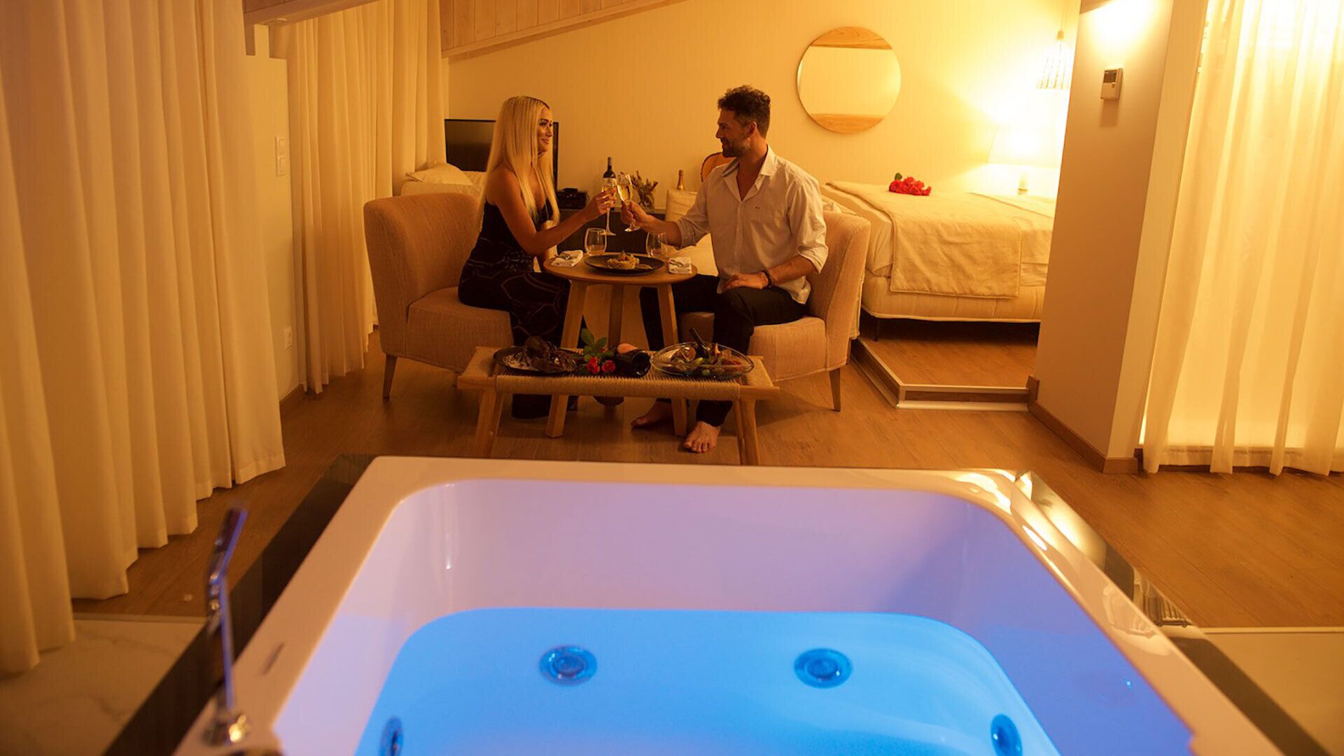 A room with a large bathtub, a dining table with food and drinks, and a bed in the background.