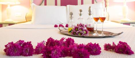 Un lit bien fait, avec du linge de lit blanc et des oreillers, un plateau avec des fleurs et des boissons, et un mur rose en arrière-plan.