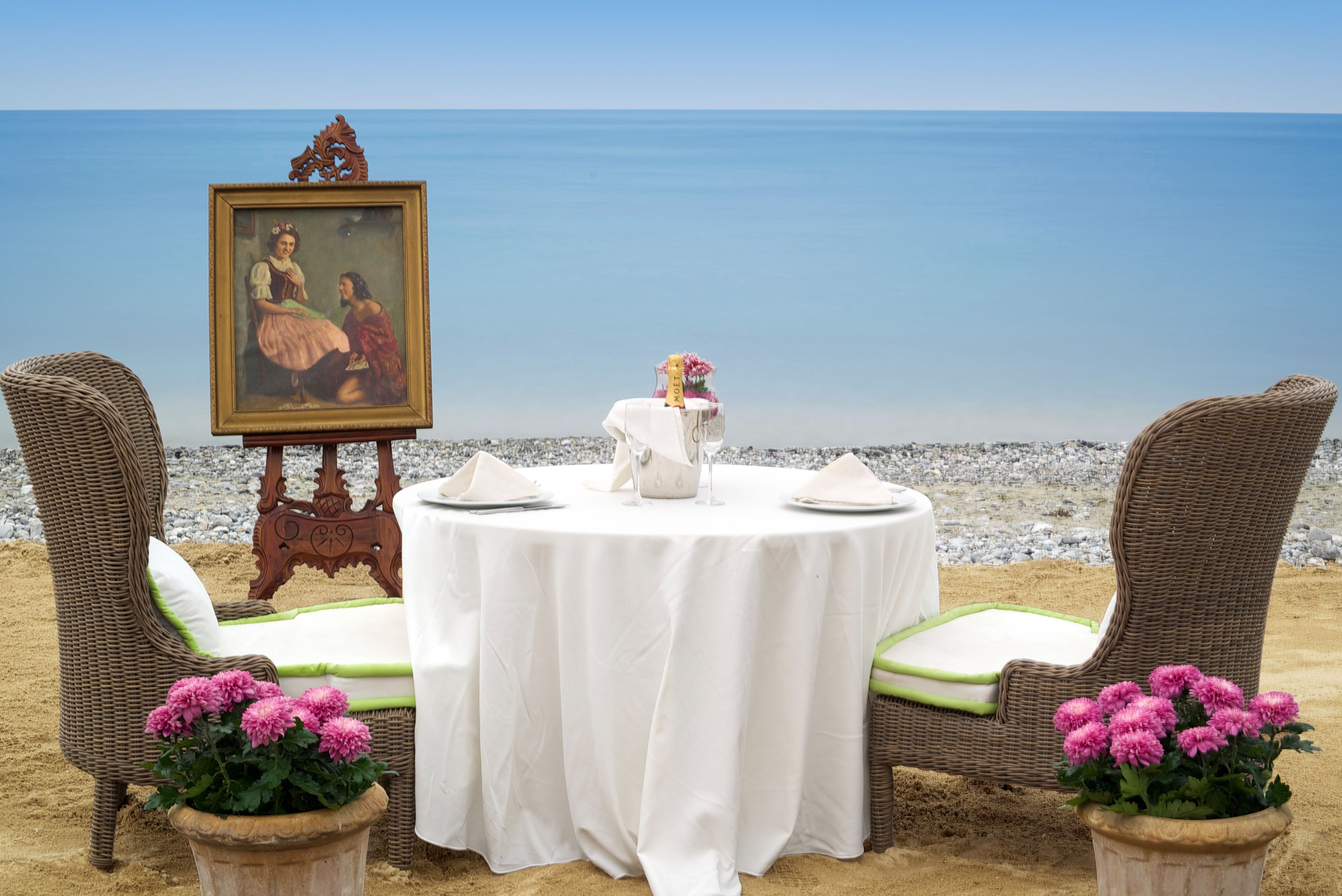 Disposição de jantar com mesa, cadeiras e um quadro apoiado, com vista para uma praia.